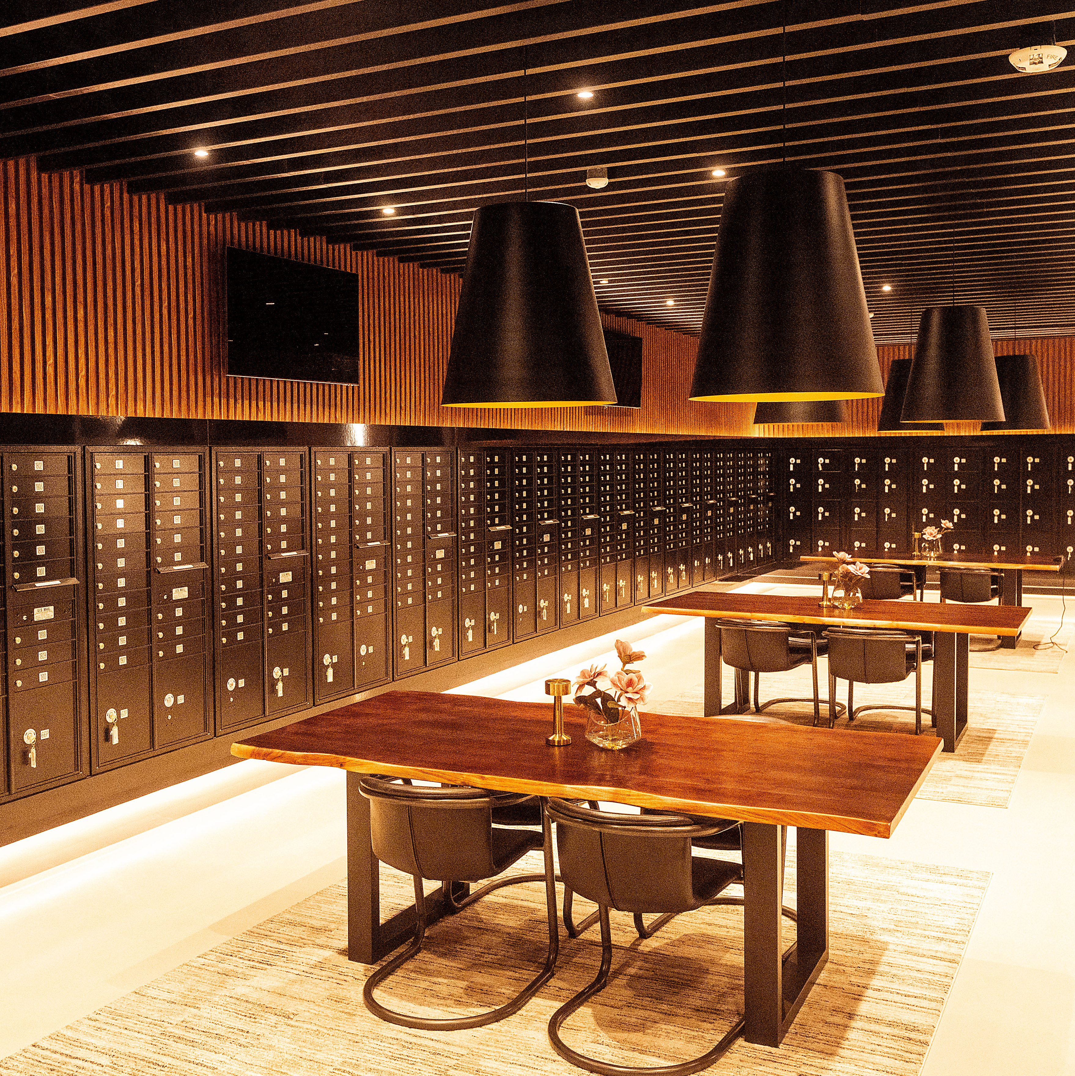 The image shows a stylish room with black lockers lining the walls, wooden tables with chairs, large black ceiling lights, and subtle warm lighting.