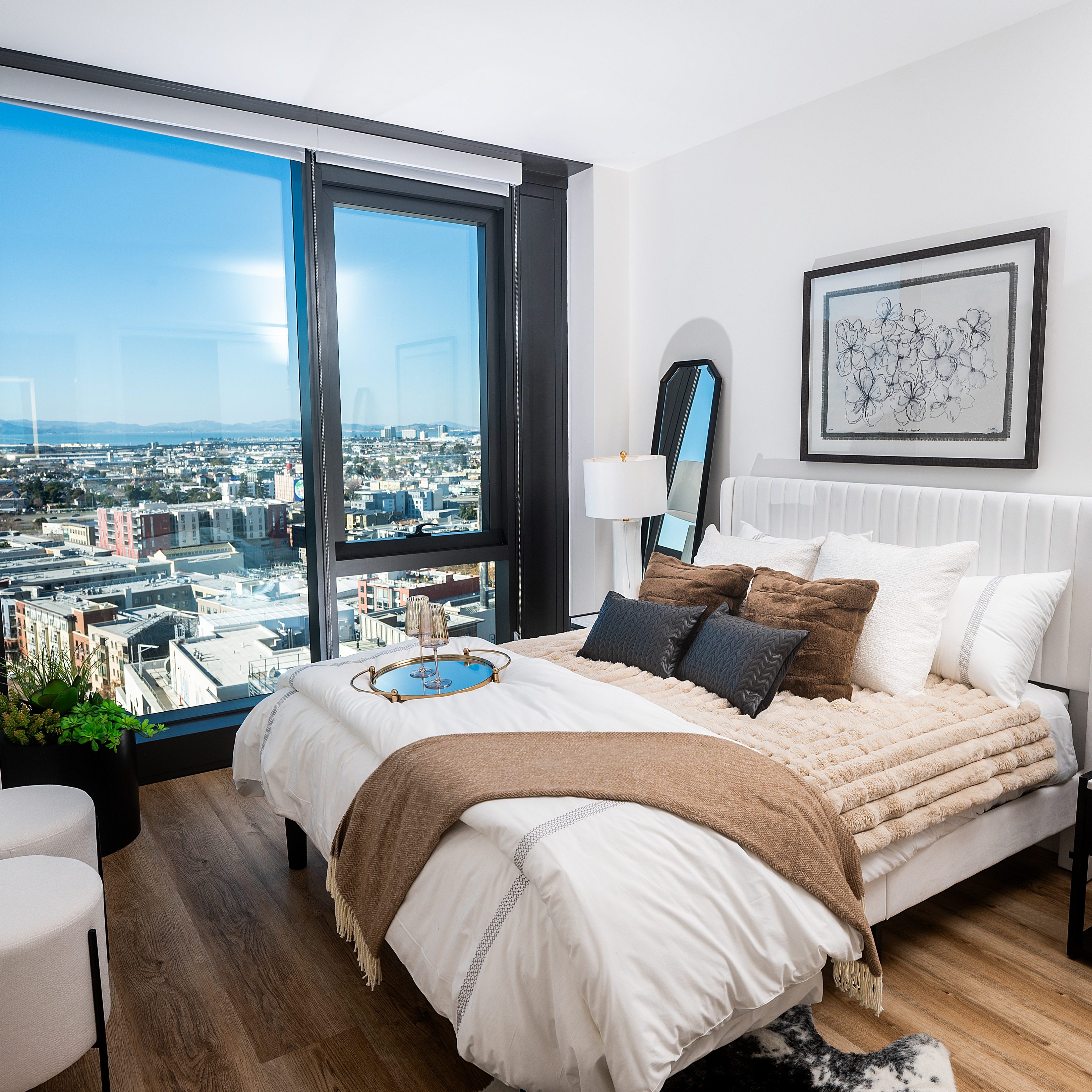 A modern bedroom features a neatly made bed with pillows and a throw. Large windows offer a city view, and there's a lamp, artwork, and plants nearby.