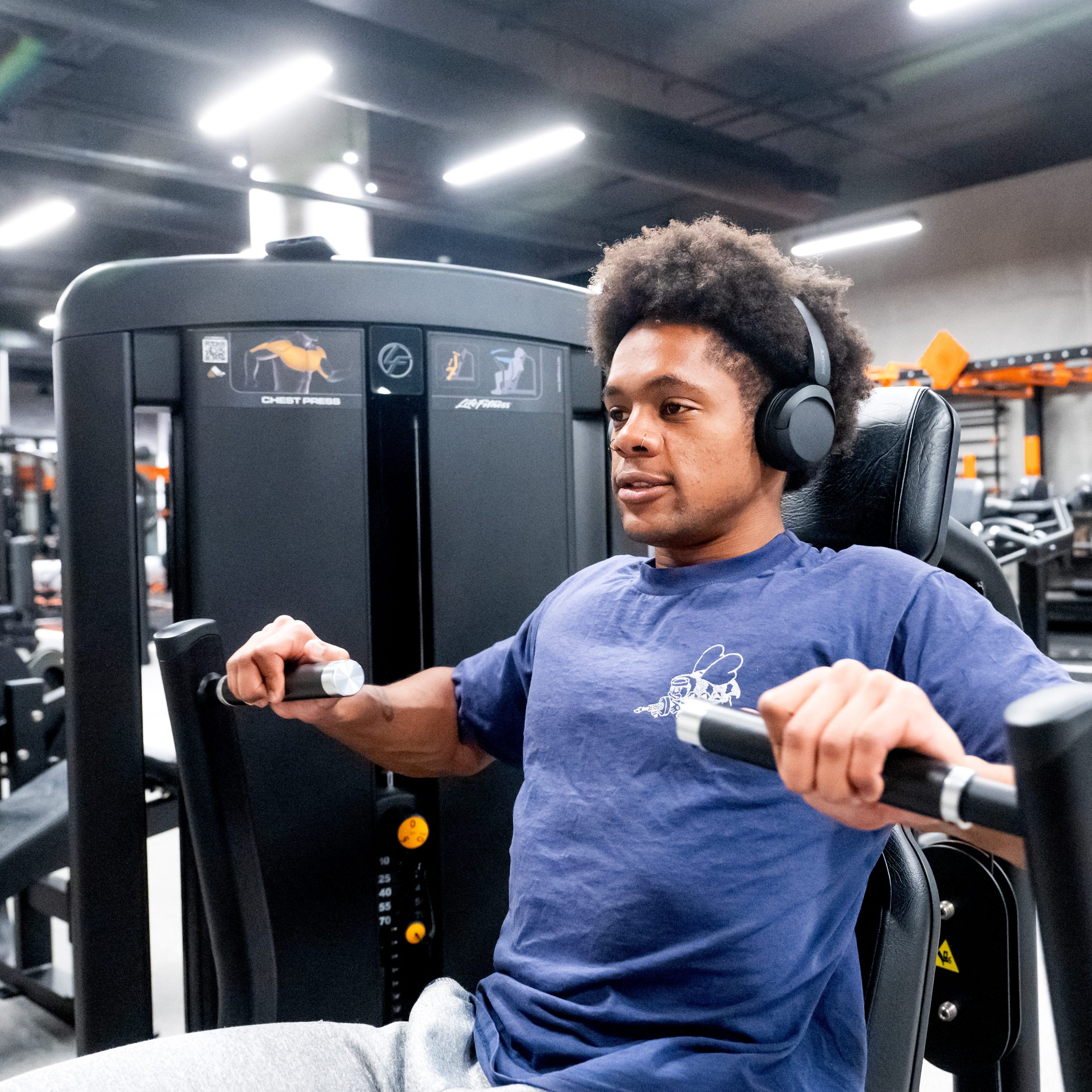 A person with headphones is using a chest press machine at the gym. They are wearing a blue shirt and appear focused on their workout.