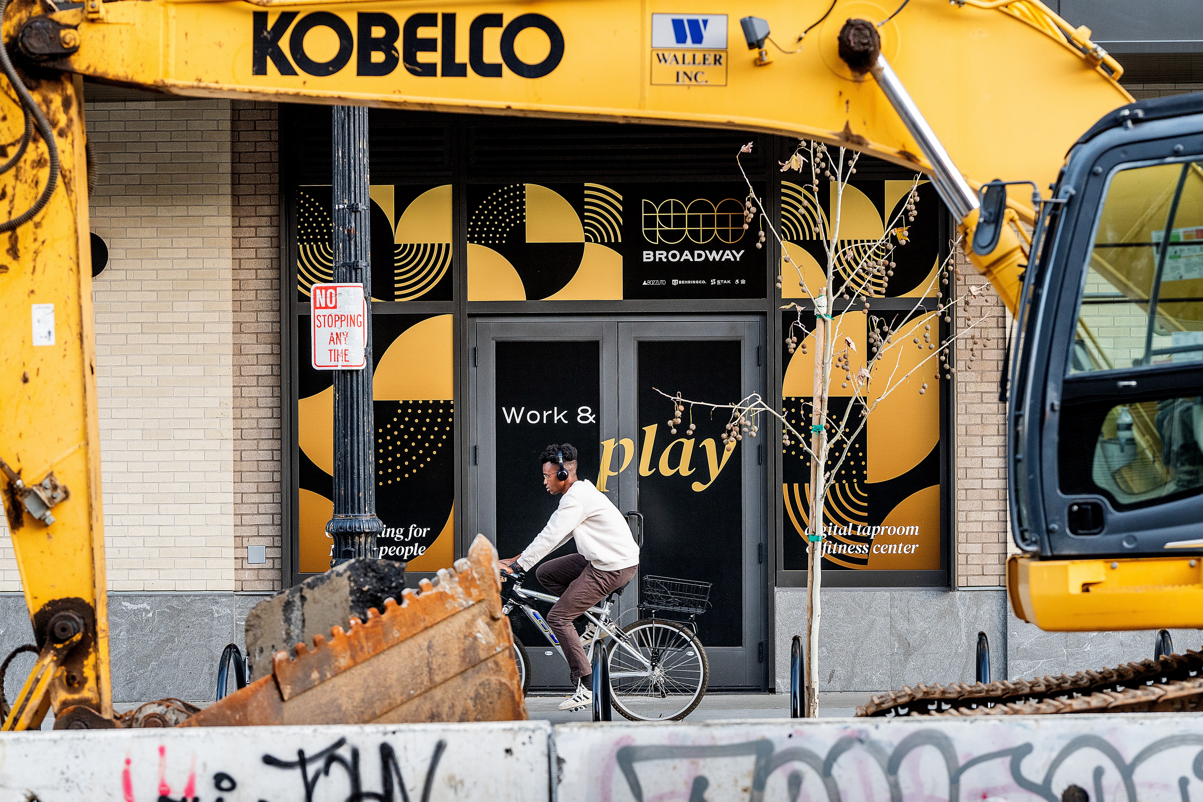 A person wearing headphones rides a bicycle between two excavators. A storefront behind them displays "Work & play" with modern yellow and black graphics.