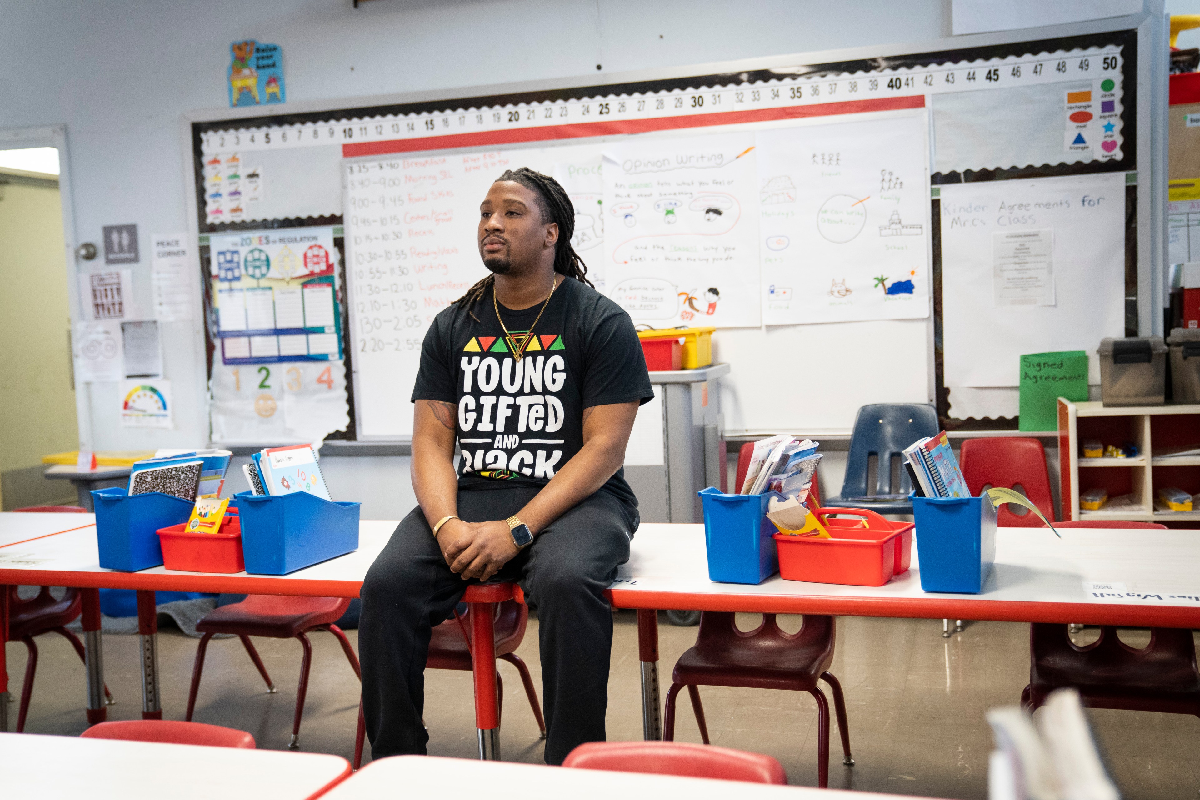 A man wearing a "Young, Gifted and Black" shirt sits on a school desk in a classroom with colorful bins and educational posters around him.