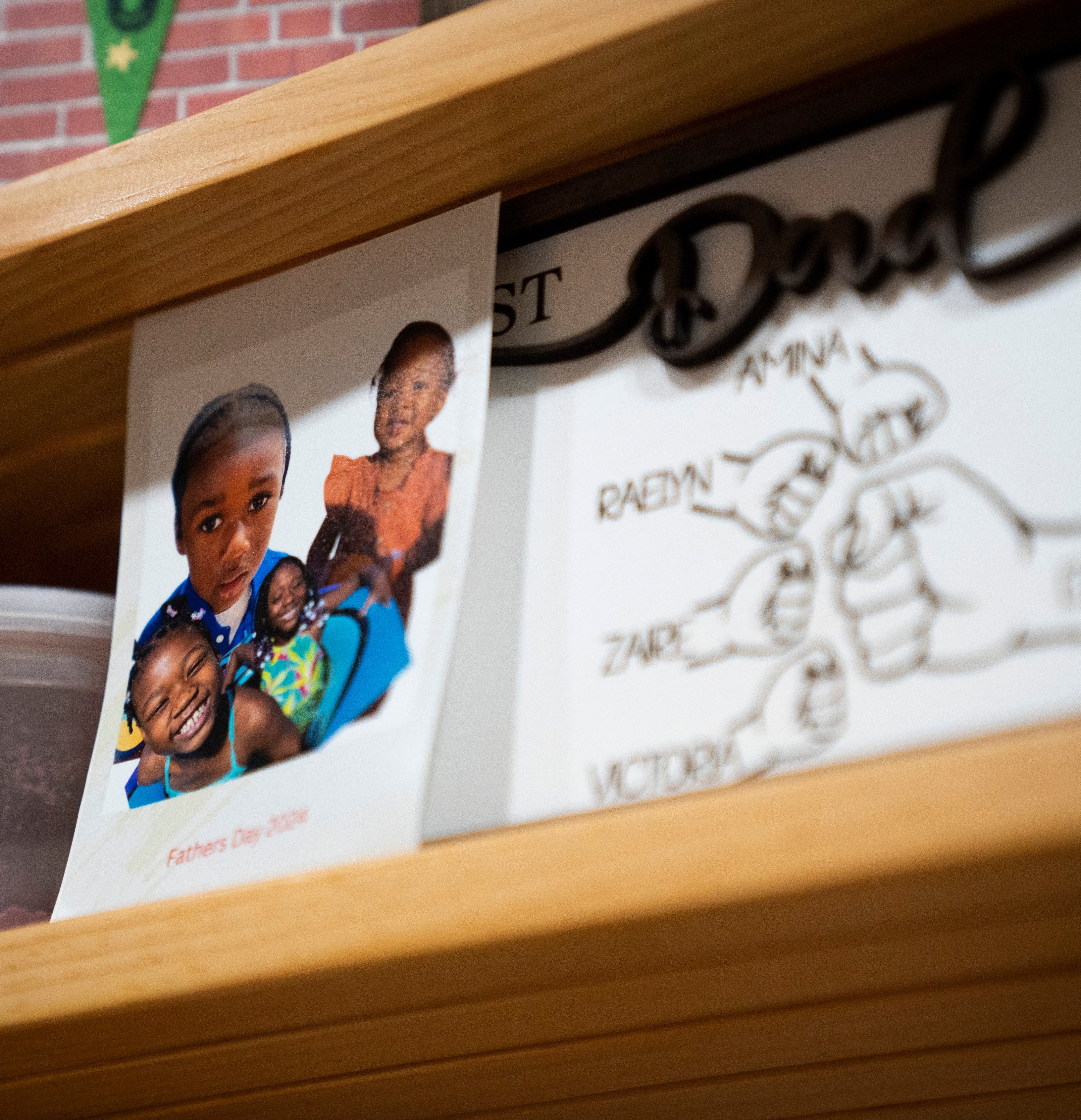 A photograph and a drawing on a wooden shelf. The photo shows smiling children with &quot;Fathers Day 2020&quot; printed below. The drawing features fists with names.