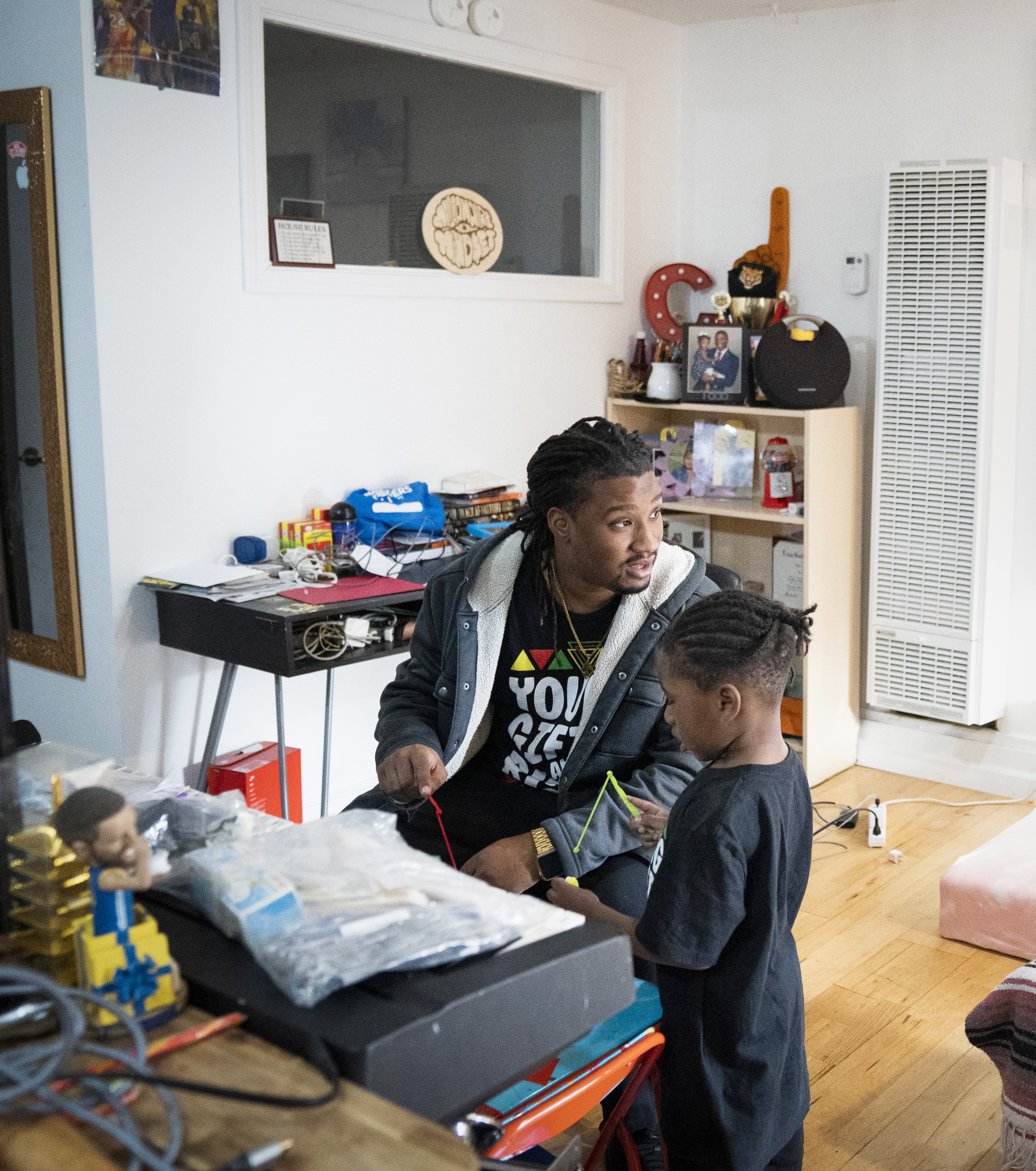 A man and a child are in a cluttered room. The man sits while the child stands, both holding items. Shelves, a heater, and various objects surround them.