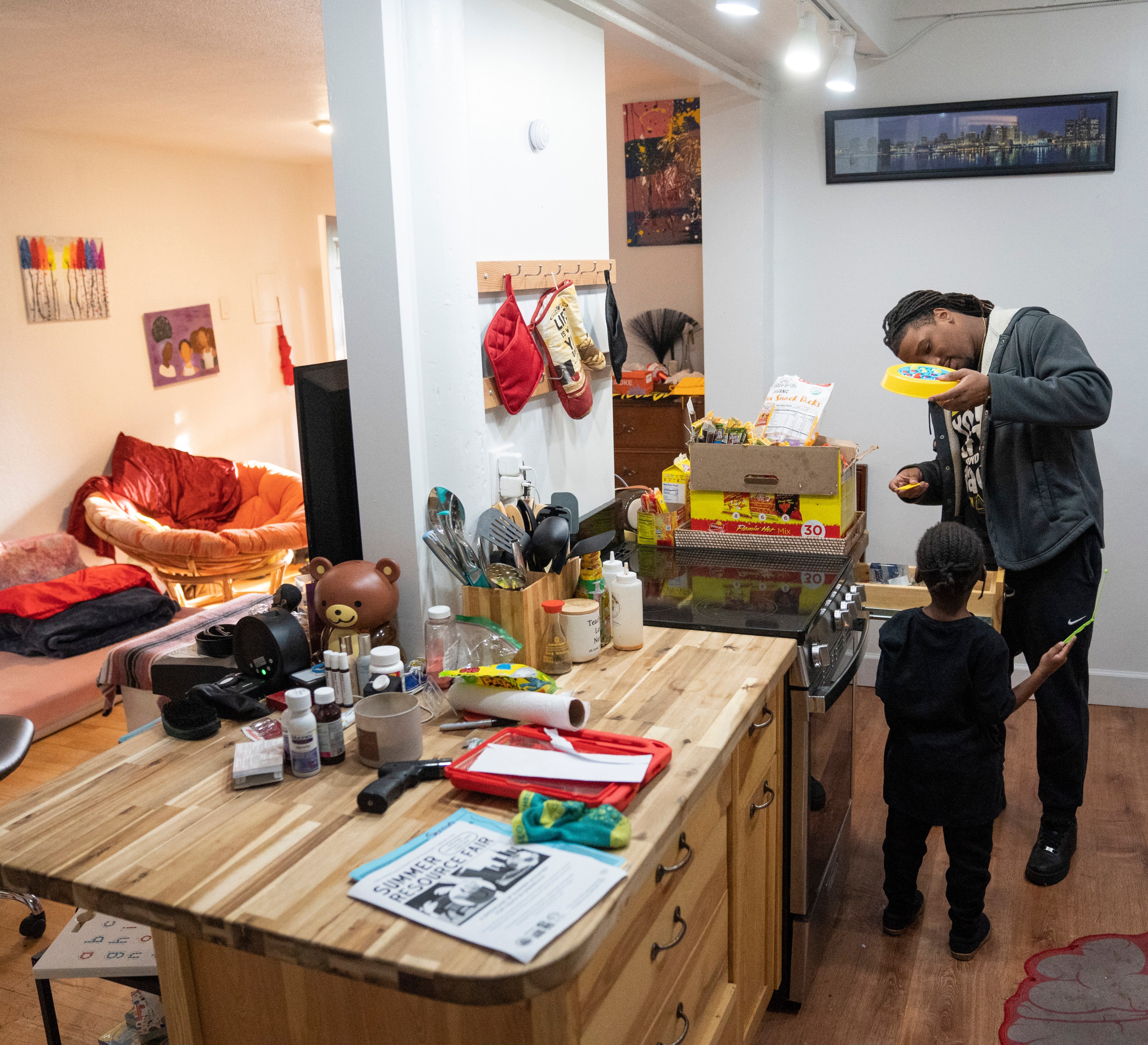 A man and child are in a kitchen; the man is holding a plate of food while the child stands near him. A cozy living area with a couch is visible.