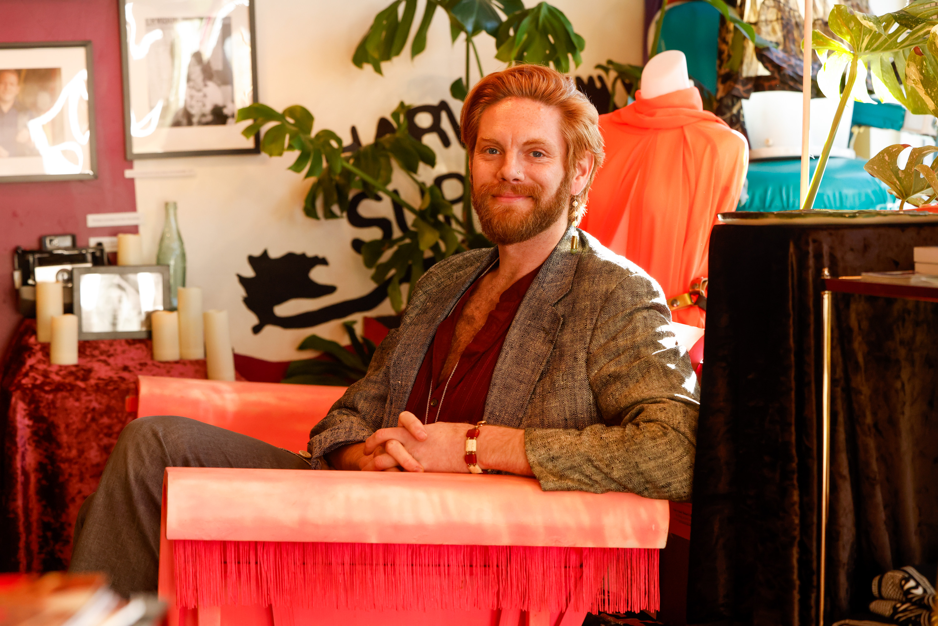 A bearded person with red hair sits relaxed in a bright pink chair, surrounded by plants and art, wearing a blazer and burgundy shirt.