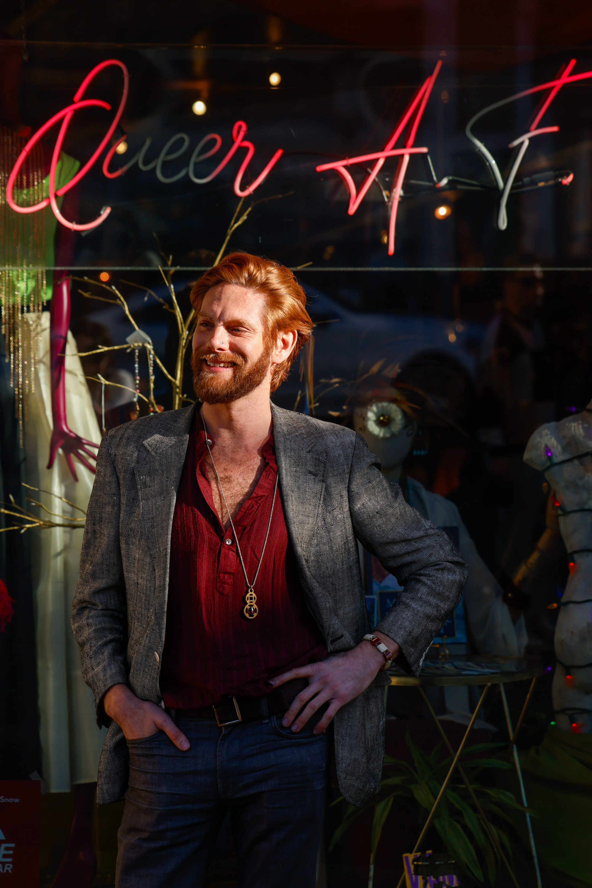 A person with red hair and a beard smiles while standing in front of a window display with mannequins and a neon sign reading "Queer AF."