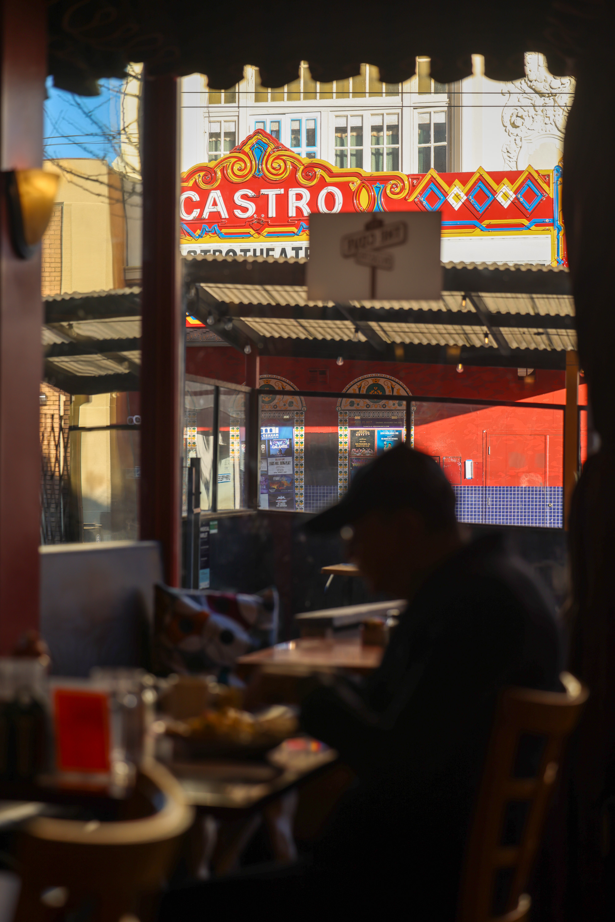 A person sits in a dimly lit cafe with a view of the colorful Castro Theatre sign outside, framed by windows and a street awning.