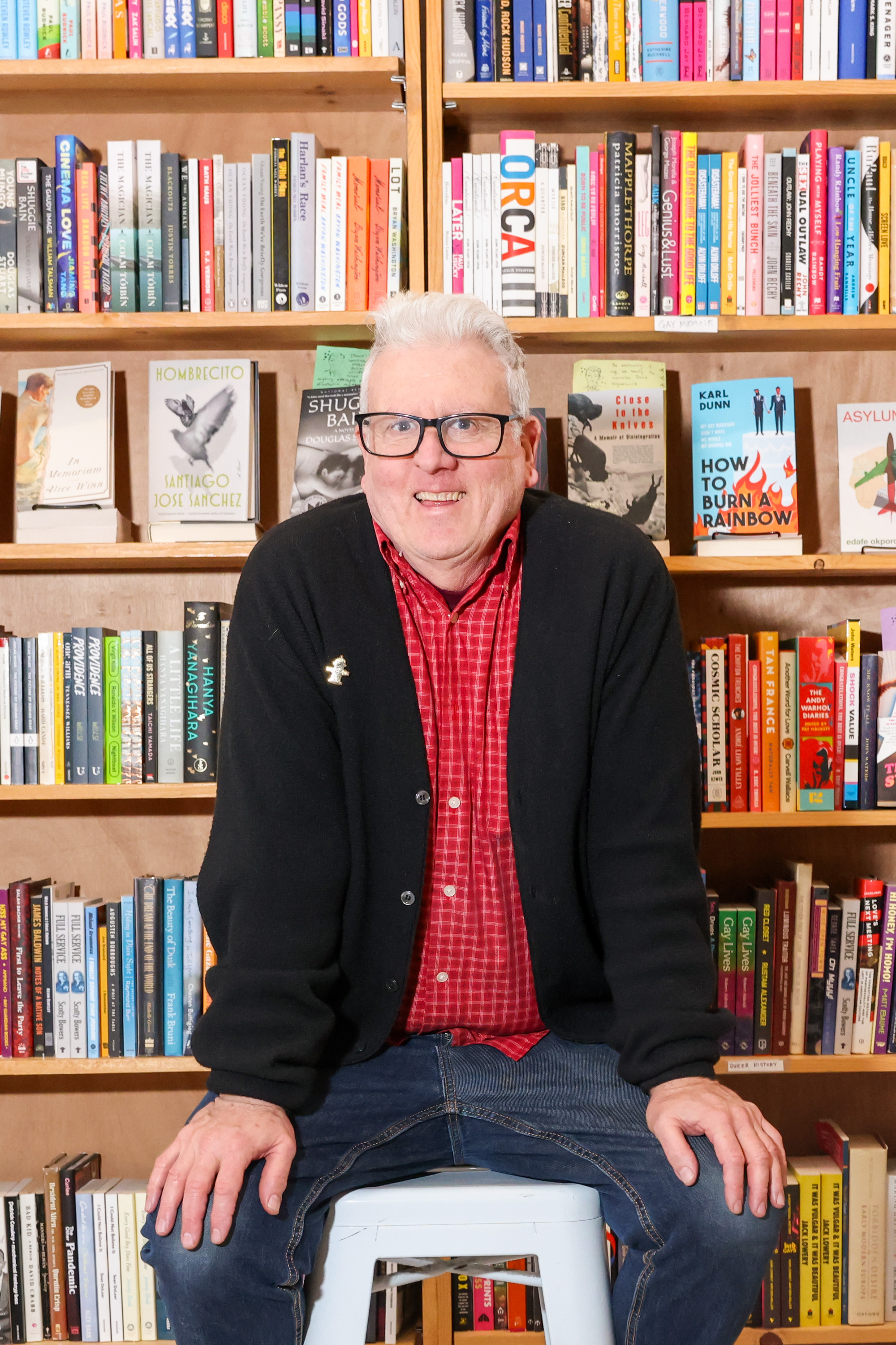 A person wearing glasses, a red checkered shirt, and a black cardigan smiles while sitting on a stool. They are in front of a colorful bookshelf full of books.