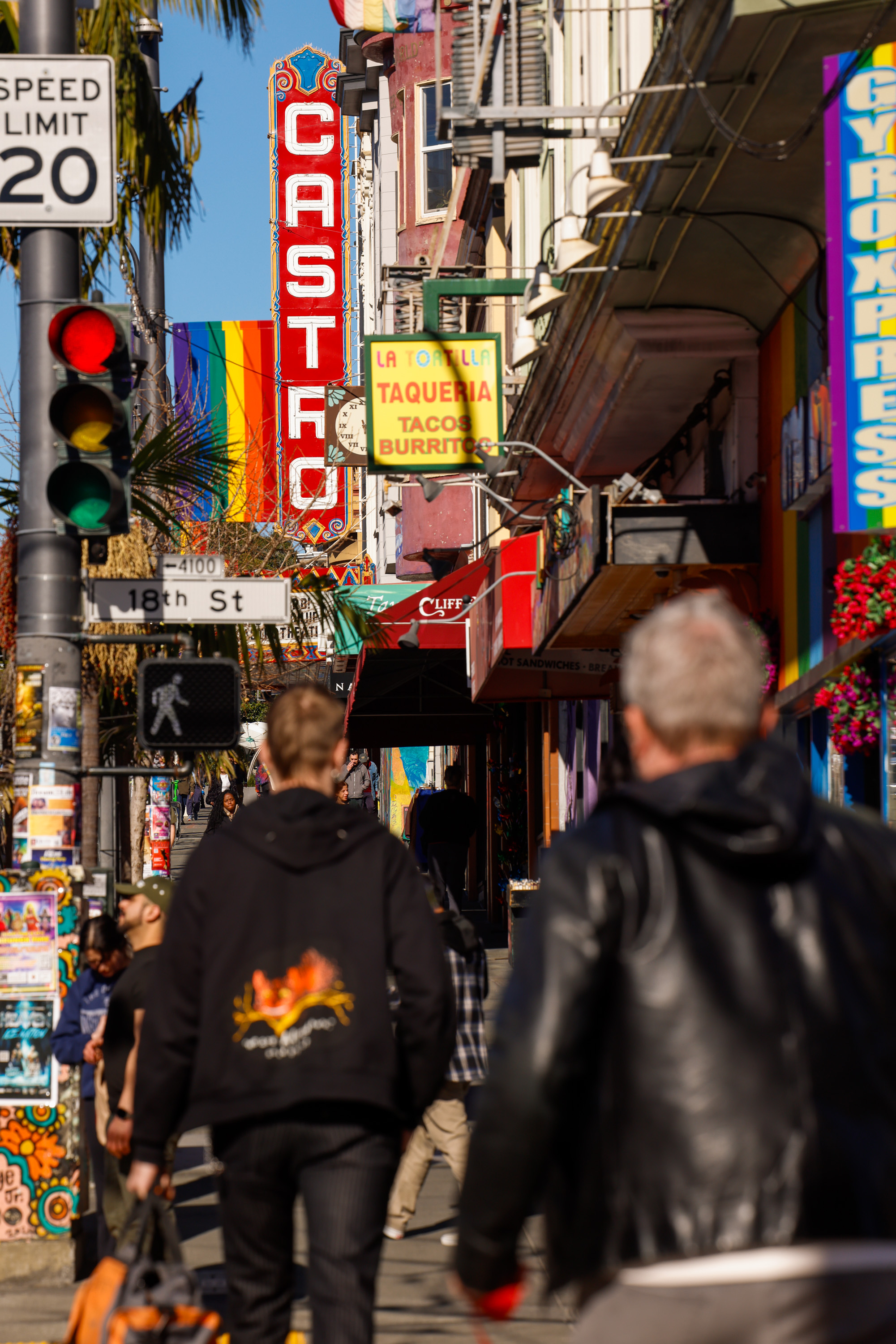 People walk along a vibrant street with colorful signs, including a large &quot;Castro&quot; sign and rainbow flag, surrounded by bustling shops and a traffic light.