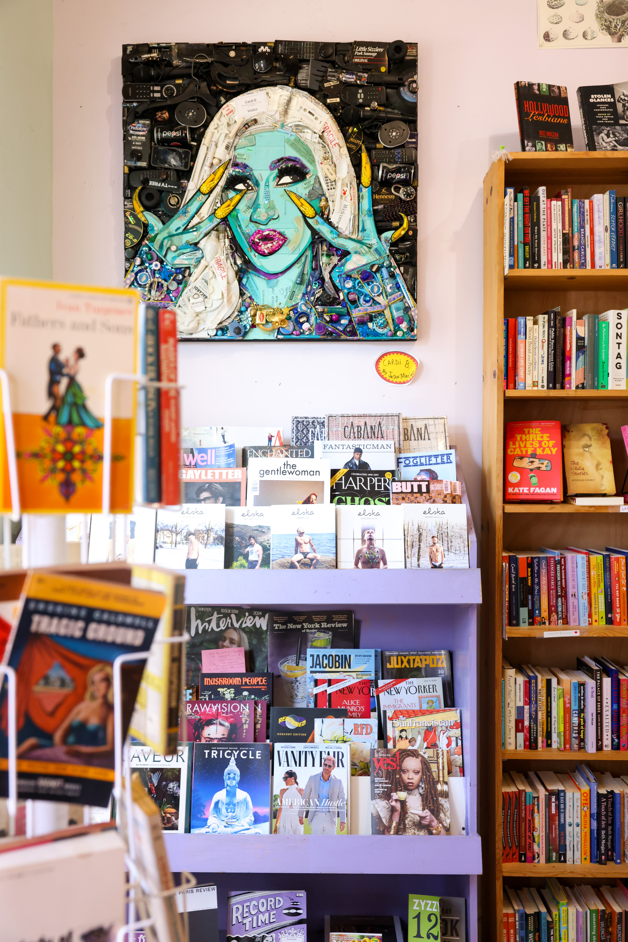 A colorful artwork of a woman's face hangs above shelves filled with books and magazines. A nearby rack holds various paperback books.