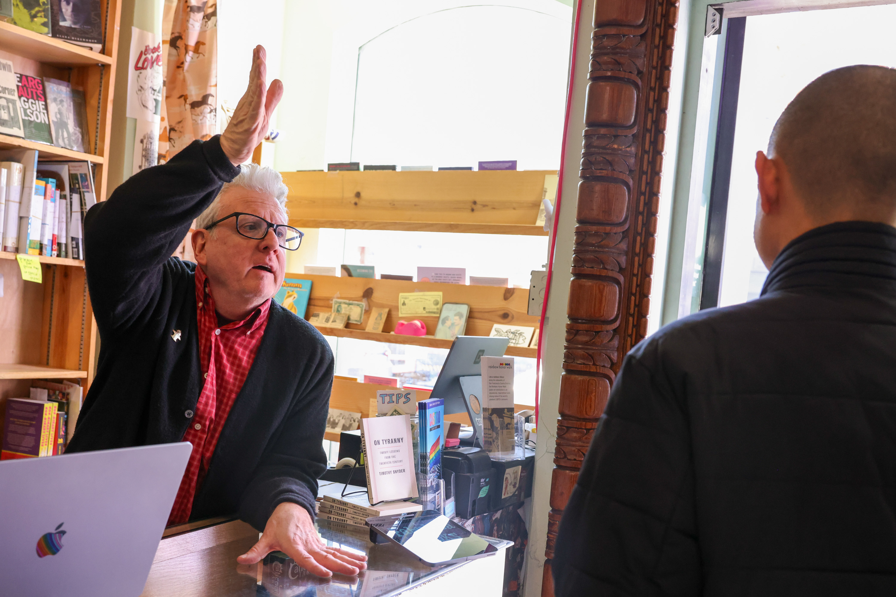 An older person with white hair, wearing glasses and a cardigan, gestures animatedly behind a bookstore counter with books and a laptop, facing a customer.