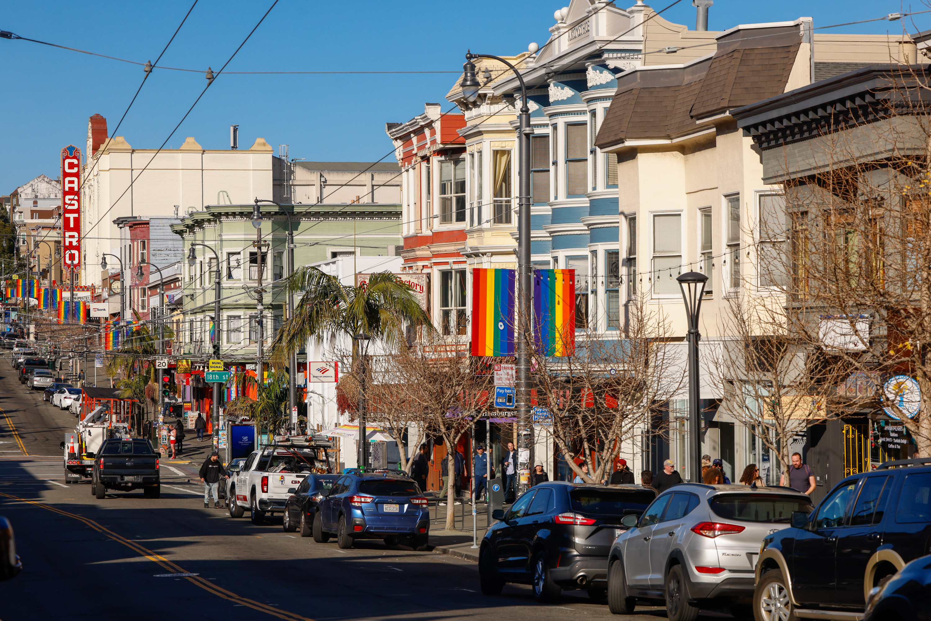 A lively street lined with colorful buildings and rainbow flags, full of parked cars and pedestrians. A large “Castro” sign is visible in the background.