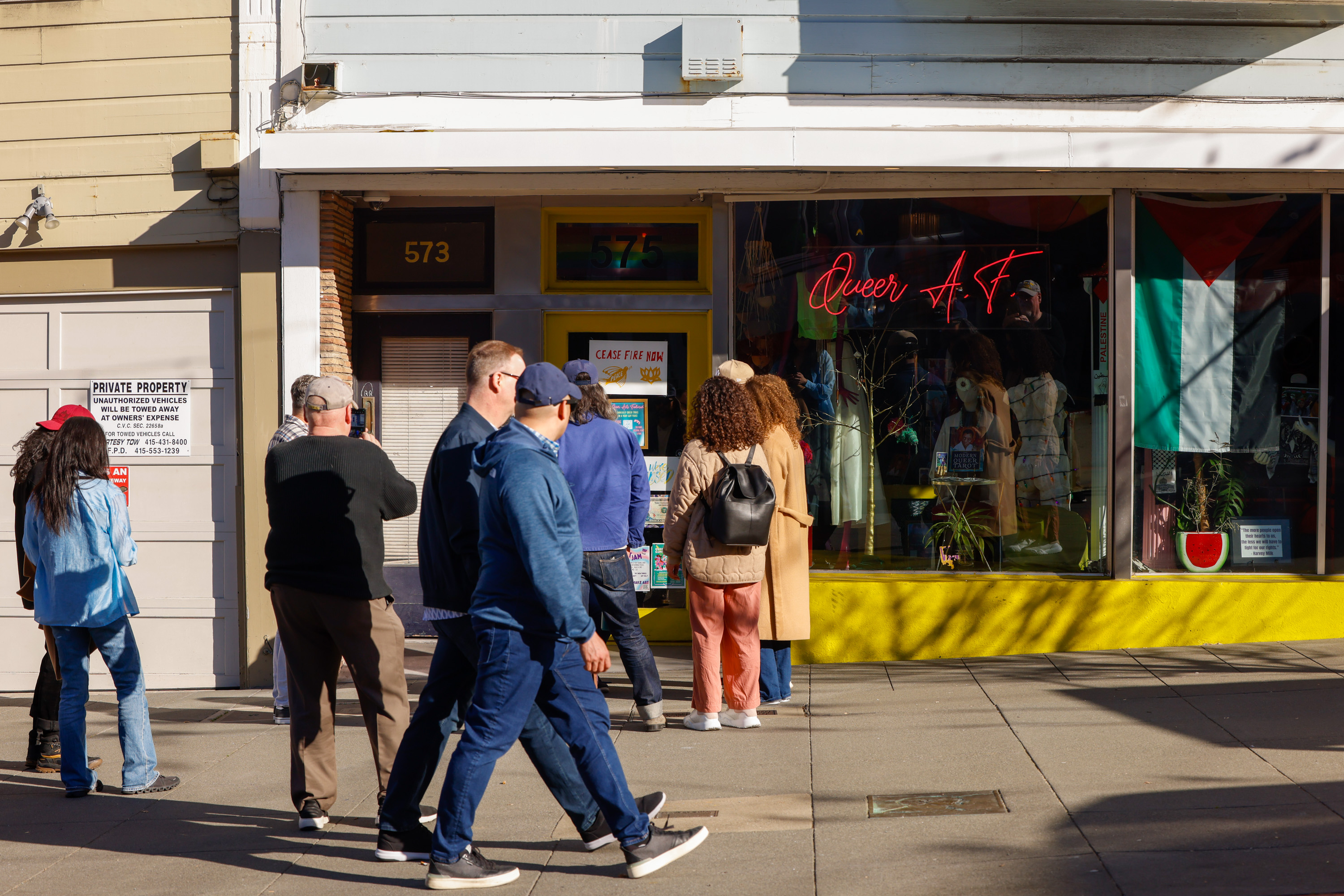 A group of people walk past a storefront with a bright yellow facade displaying a &quot;Queer A.F&quot; neon sign and various colorful items in the window.