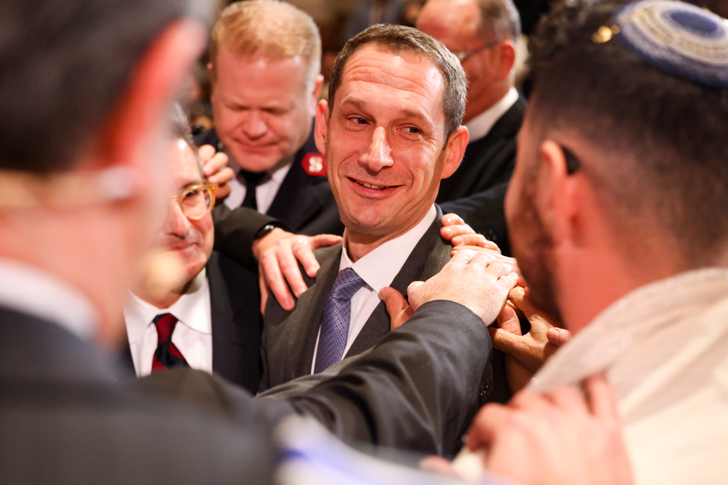 A group of people, mostly men in suits, gather closely, smiling and touching a central figure's shoulders in a supportive, celebratory manner.