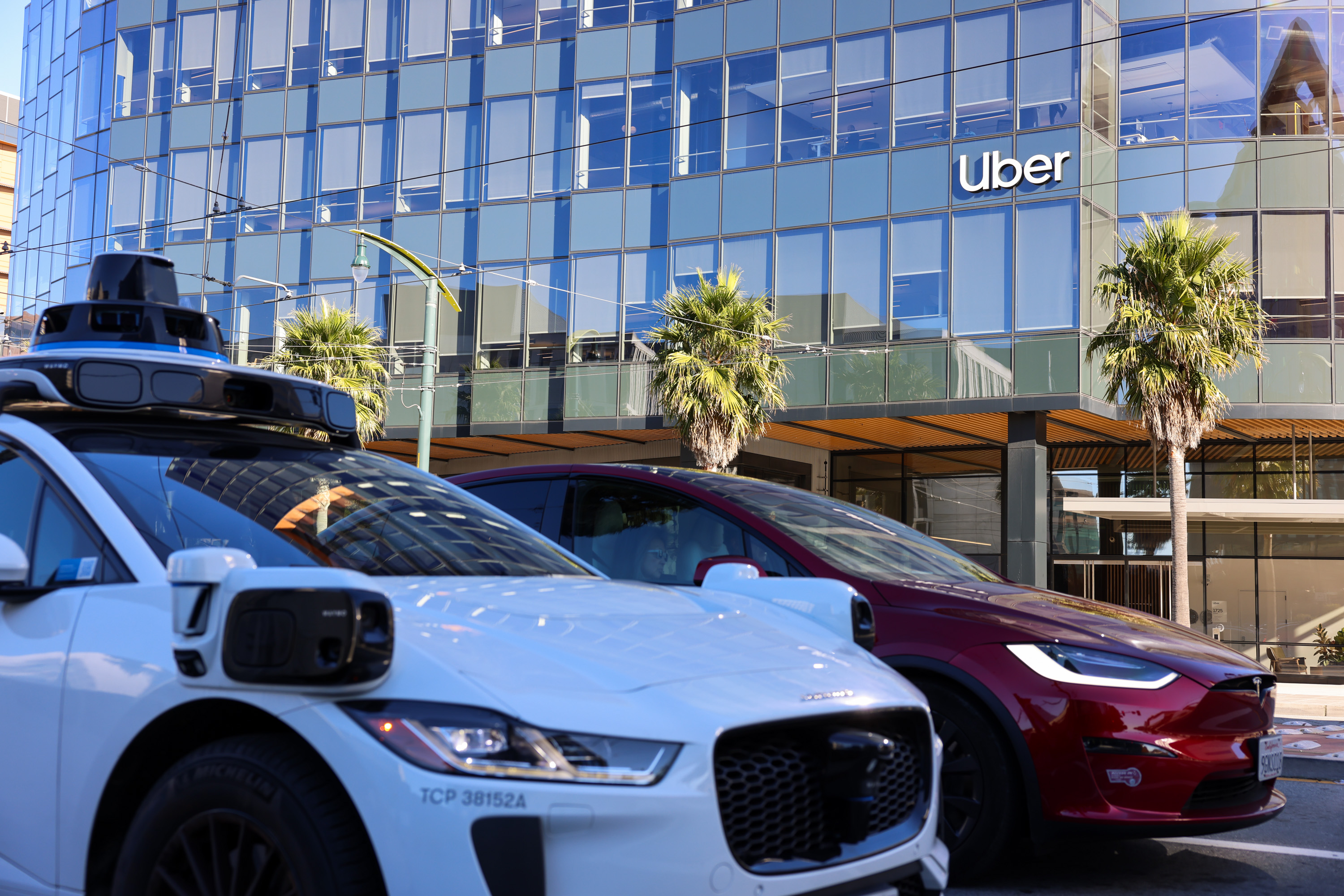 A white autonomous vehicle with sensors is parked alongside a red car in front of a modern building with the Uber logo and palm trees visible.