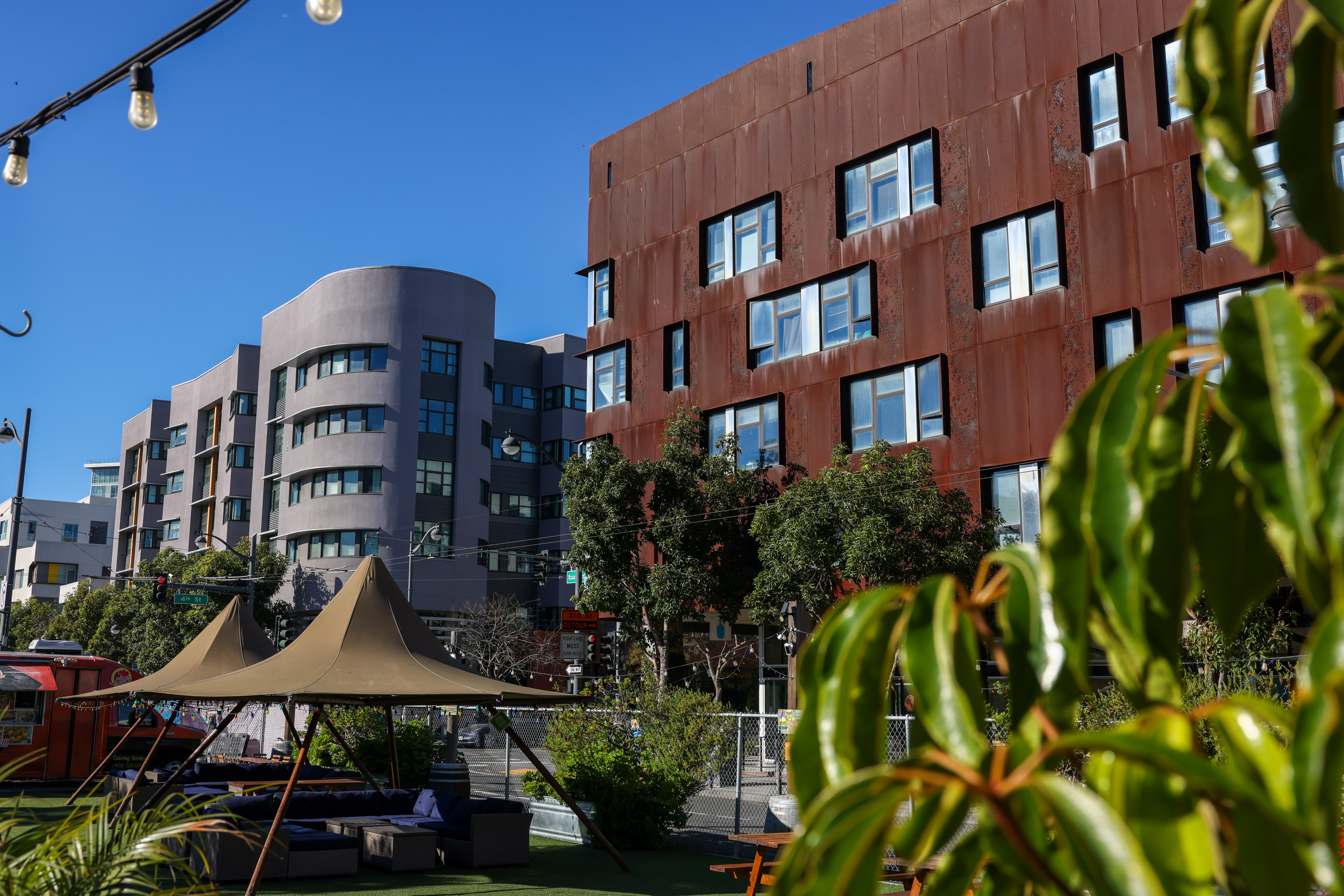 A modern urban scene features two distinctive buildings, one grey and curved, the other rust-colored. Trees, plants, and string lights add a touch of greenery.