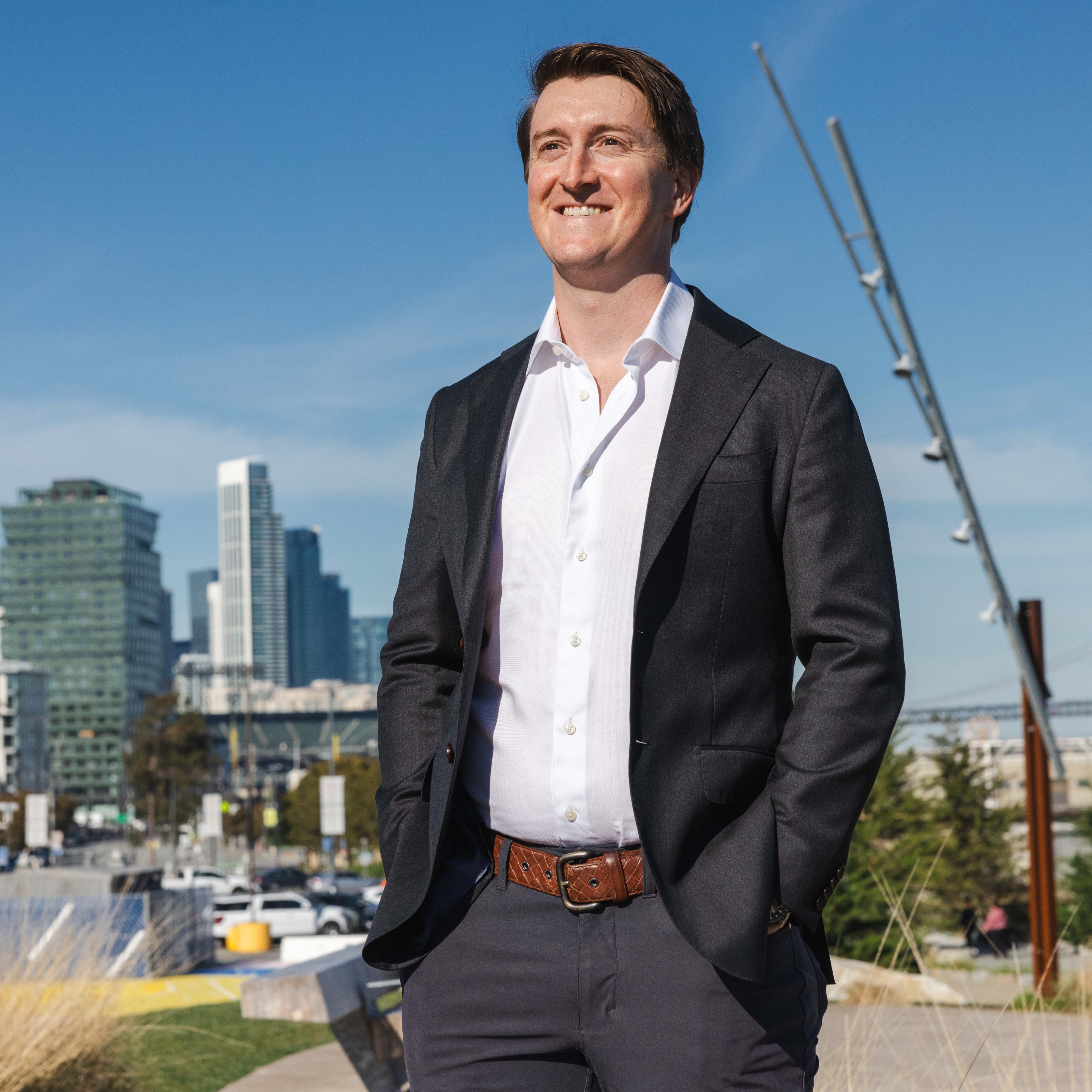 A person in a dark suit and white shirt stands outdoors smiling, with a city skyline and bridge in the background on a clear day.