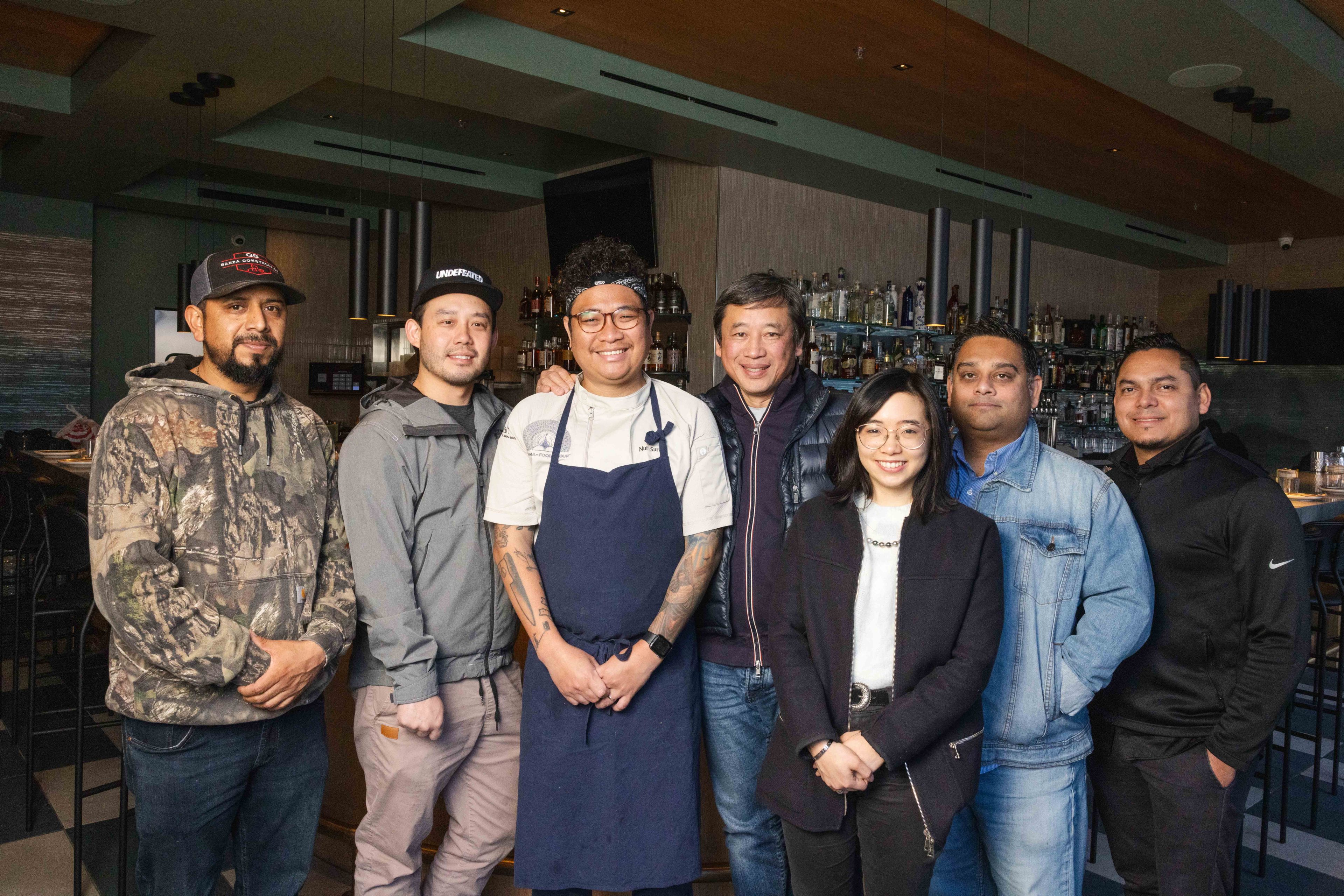 A group of seven people stands smiling in a restaurant setting. One person wears an apron, and the bar is visible behind them. The atmosphere is casual.