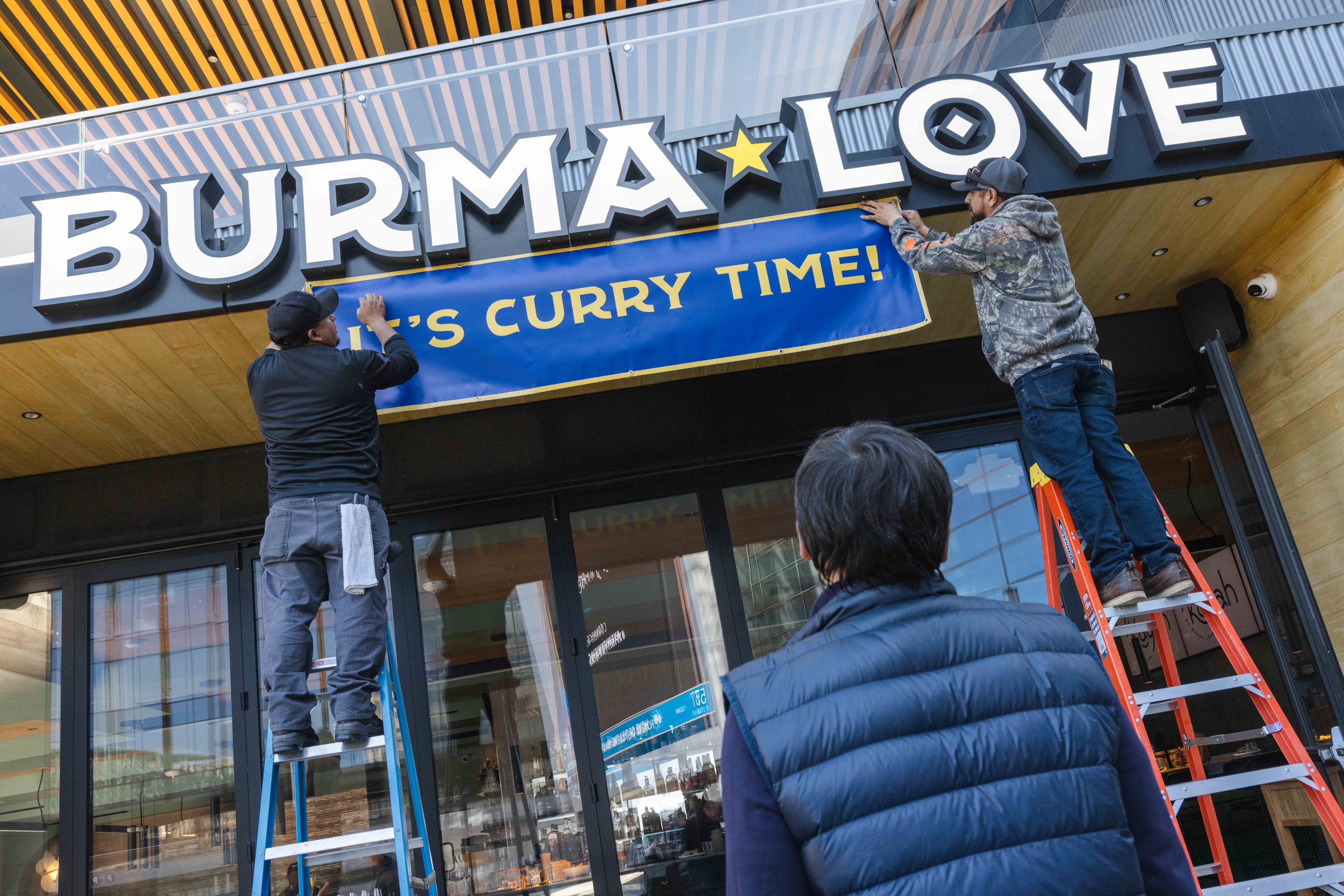 Two men on ladders are hanging a blue banner saying &quot;IT'S CURRY TIME!&quot; below a sign that reads &quot;BURMA LOVE.&quot; A person in a black vest watches.