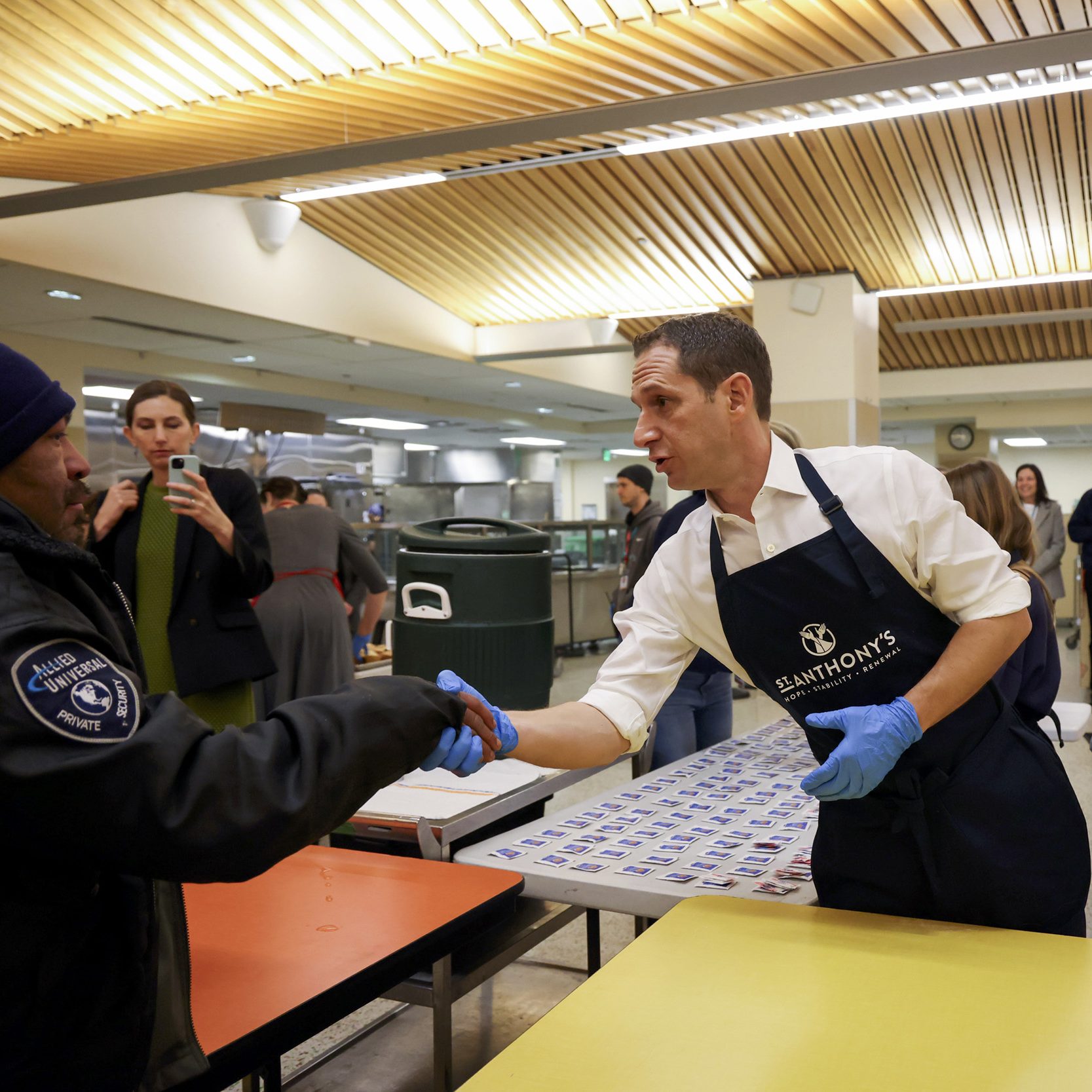 A man in a St. Anthony's apron shakes hands with another person in a communal dining area. People are gathered around, and a child stands nearby.