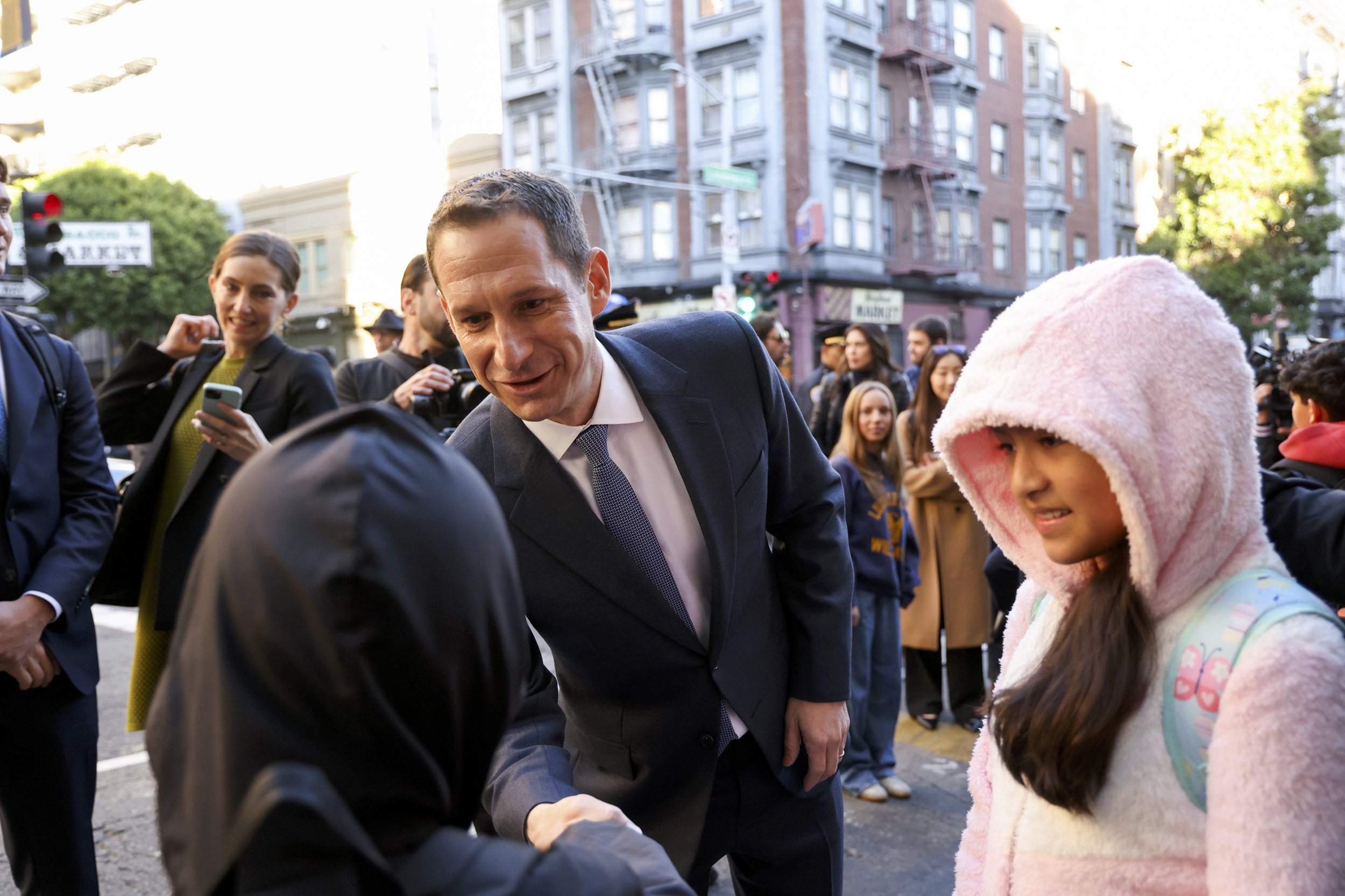 A man in a suit shakes hands with someone wearing a black hood. A girl in a pink hooded jacket smiles nearby, while onlookers, including a woman with a phone, watch.