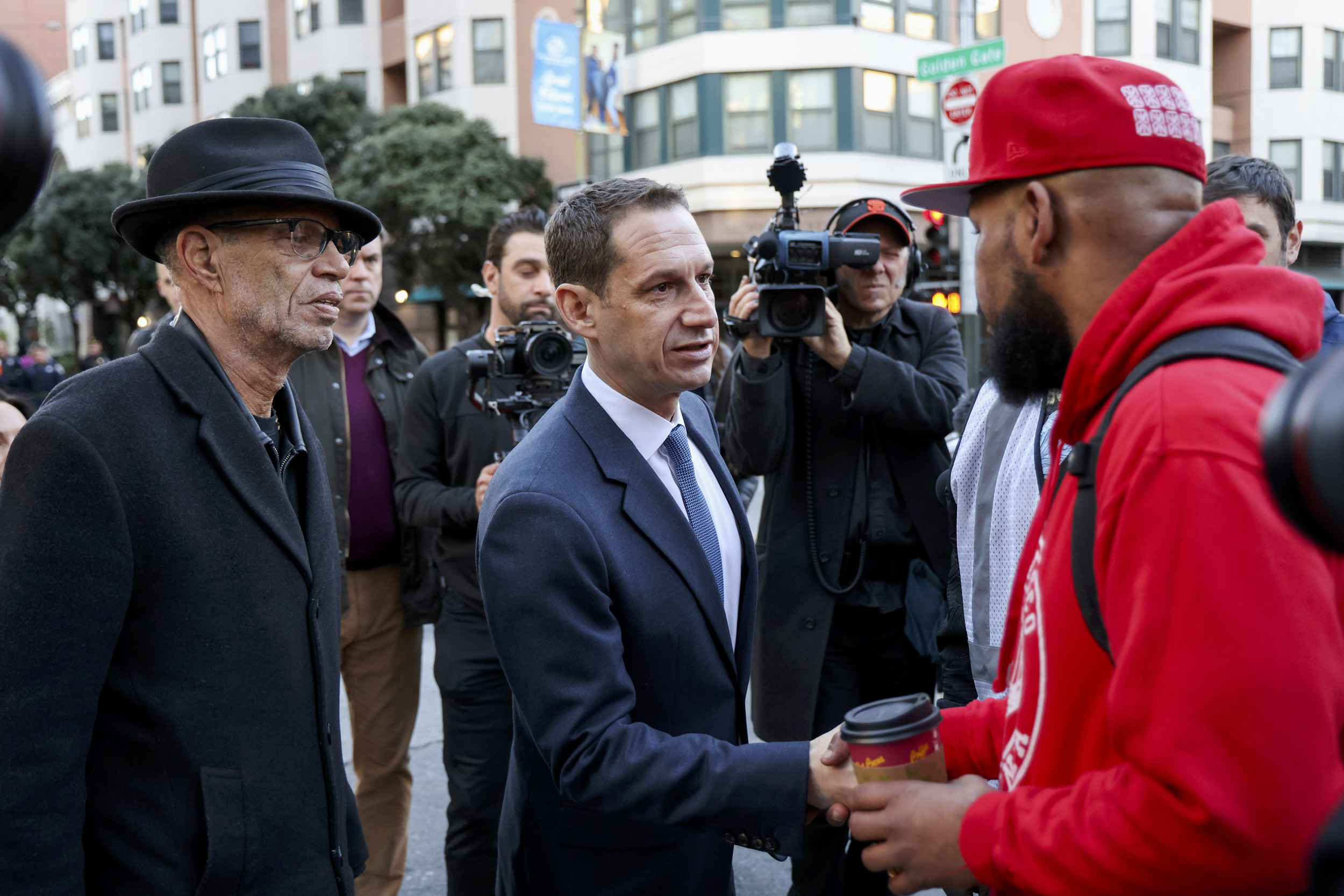 A man in a suit shakes hands with a man in a red hoodie and cap on a busy street, surrounded by people and cameras, with buildings in the background.