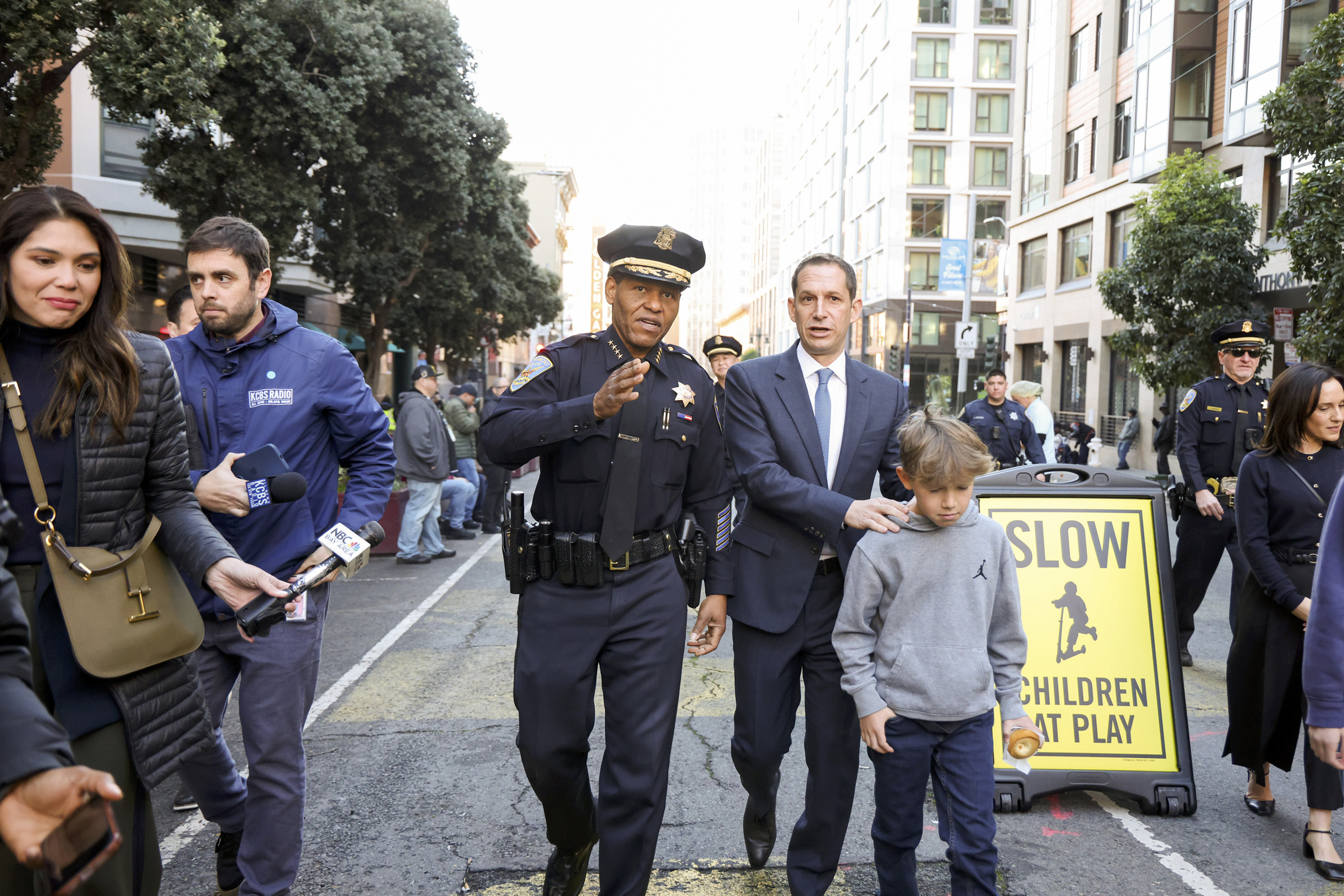 A police officer walks down a city street alongside a man in a suit and a boy. Journalists and onlookers are nearby. A "Slow Children at Play" sign is visible.