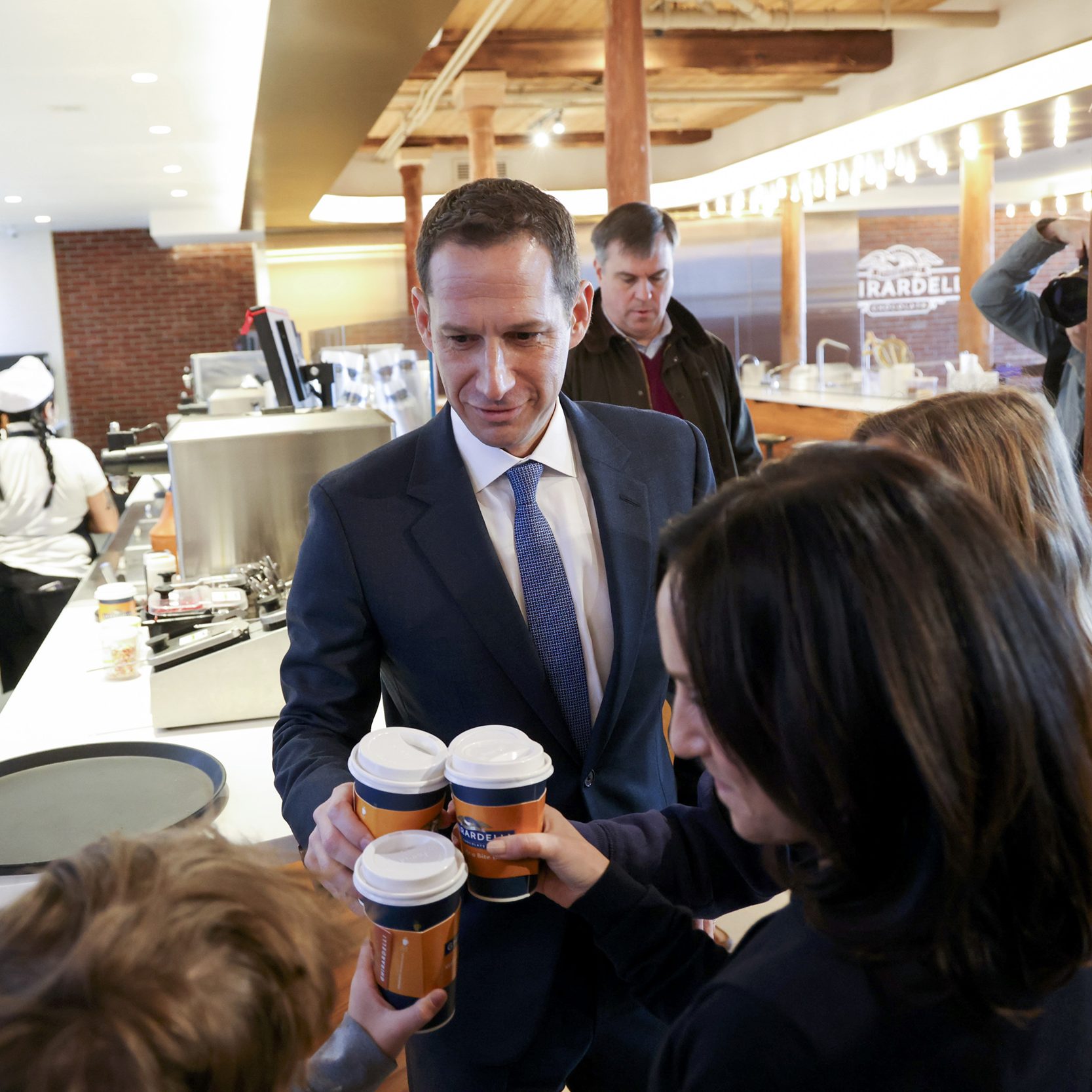 A man in a suit hands out coffee cups to people in a cafe, while staff in aprons work behind the counter. Photographers capture the scene.