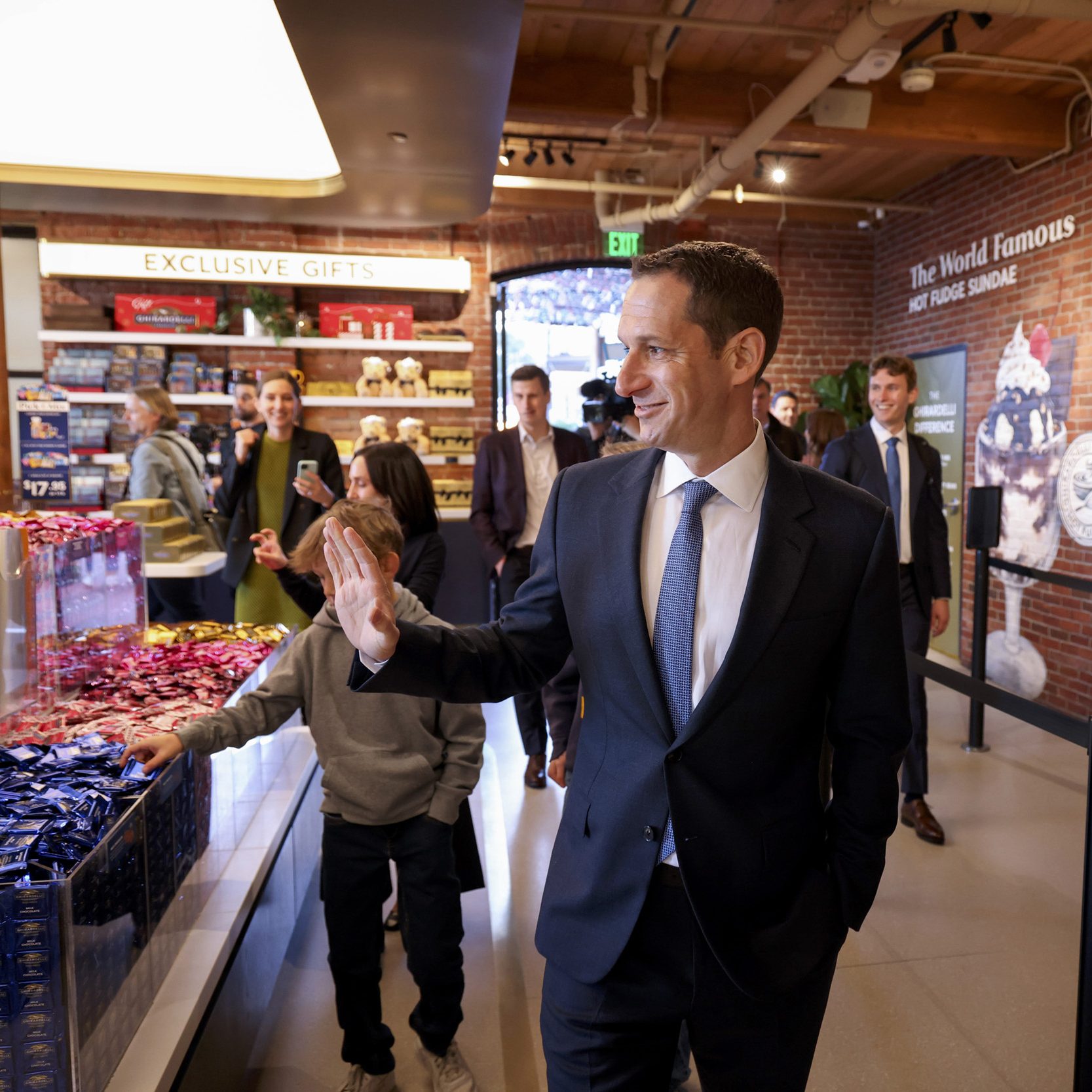 A man in a suit waves in a shop filled with candy. A child reaches for chocolate, while patrons and staff stand around and an &quot;Exclusive Gifts&quot; sign is visible.