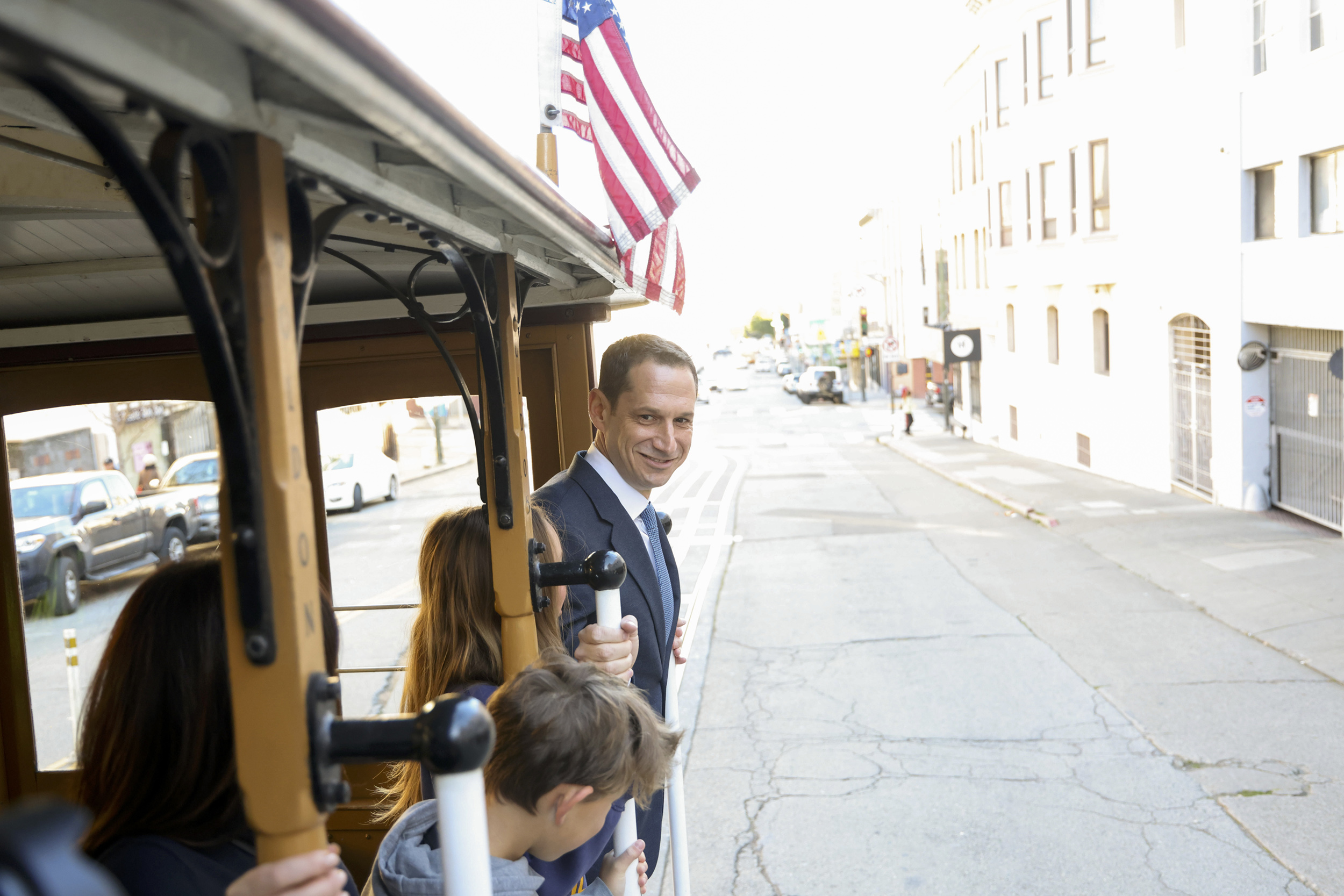 A man in a suit smiles while riding a streetcar, standing near a boy. There's an American flag above them, and the street is lined with buildings and parked cars.