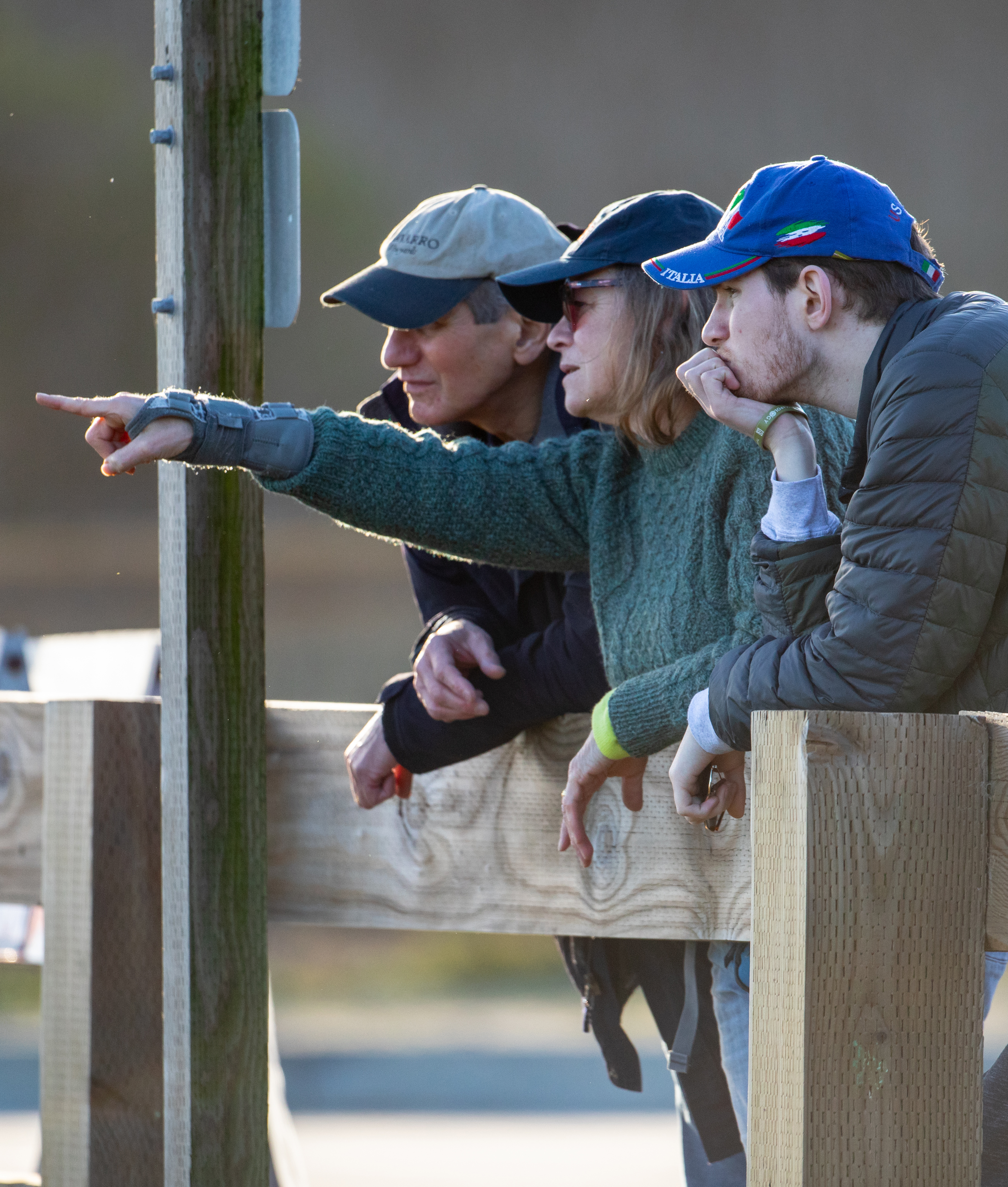 Three people are leaning on a wooden fence, looking intently in the same direction. One person is pointing, all are wearing caps, and dressed warmly.