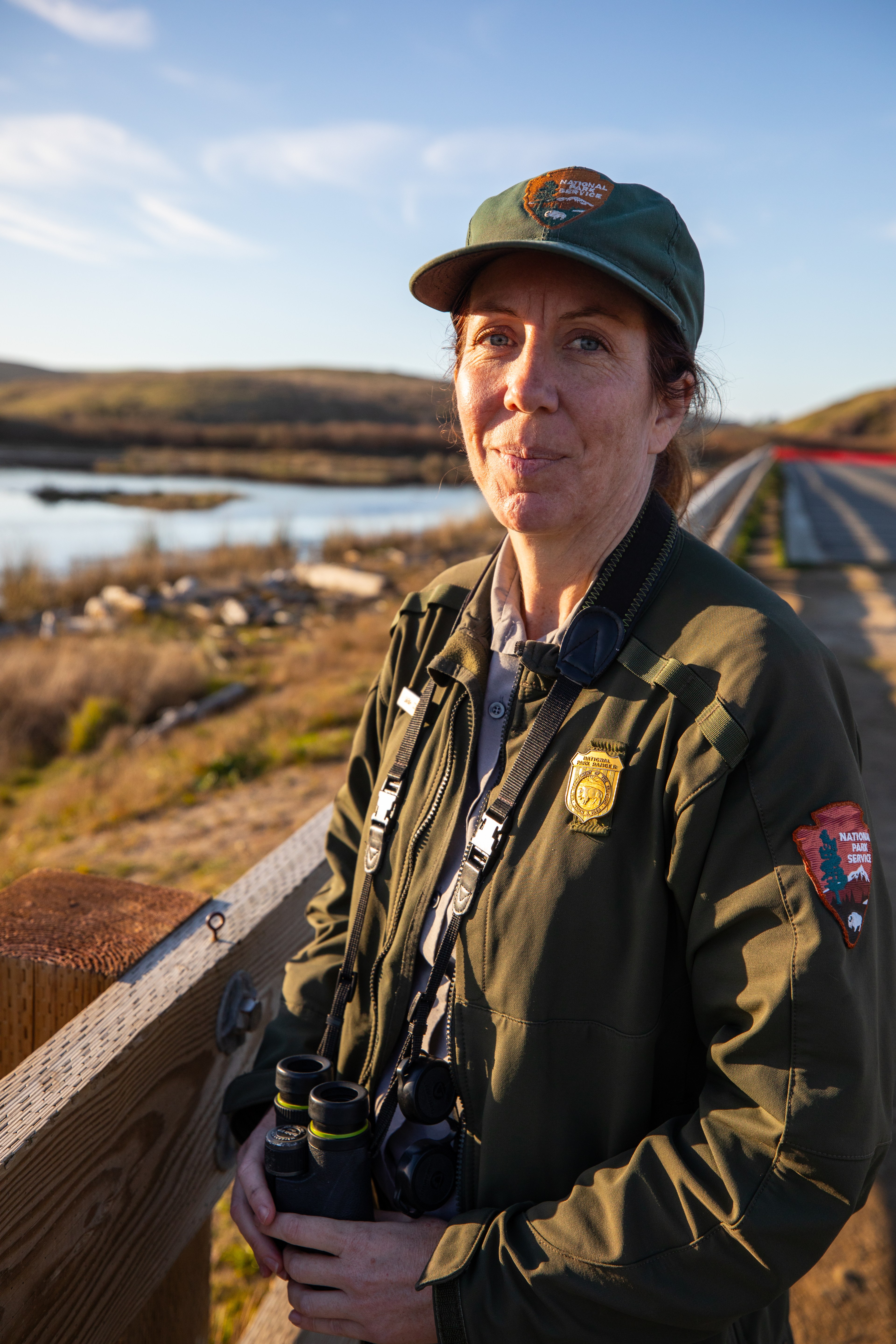 A park ranger in uniform stands beside a wooden railing, holding binoculars. She is outdoors with a scenic background of hills and a body of water under a clear sky.