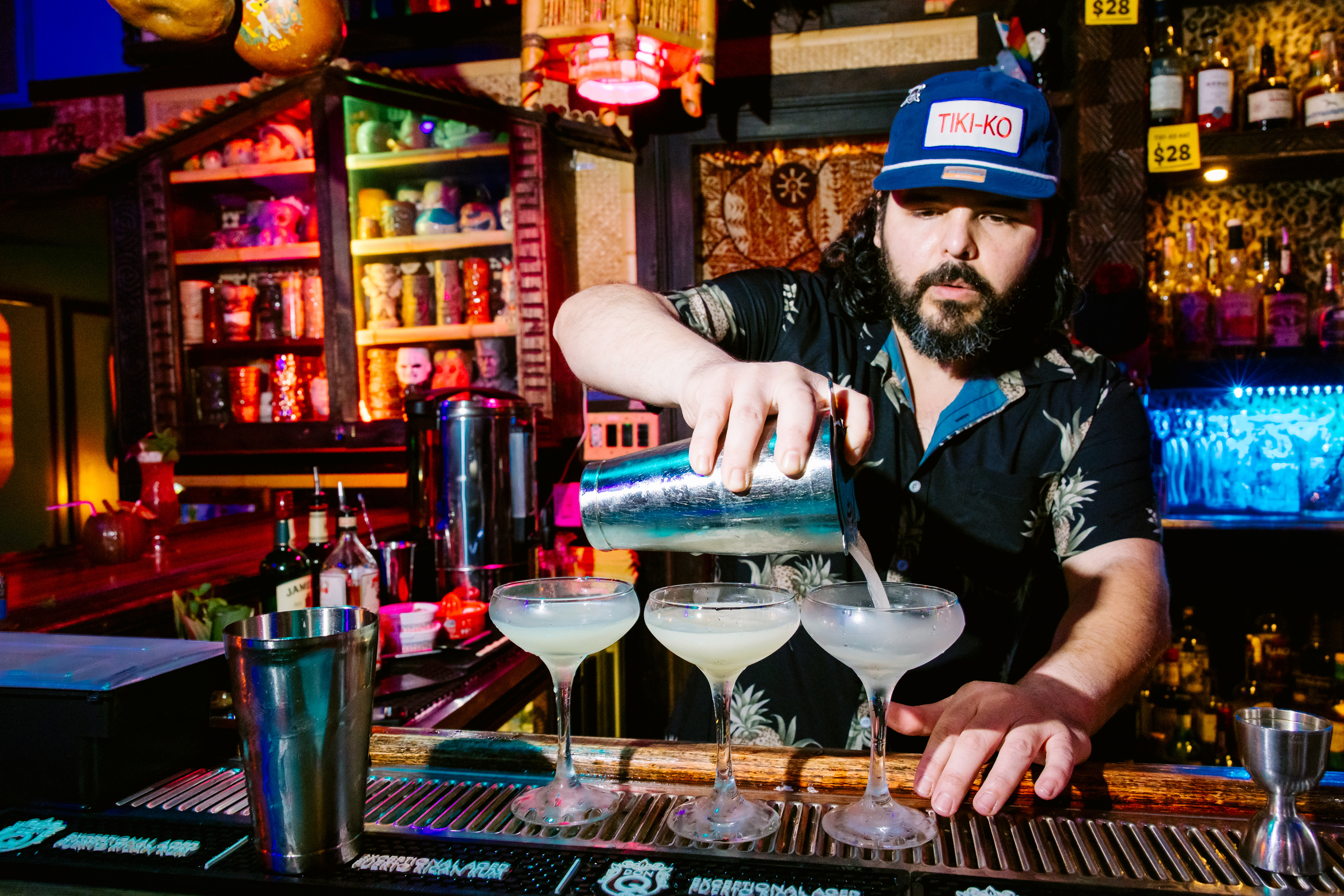 A bartender in a floral shirt and "TIKI-KO" cap pours drinks into three cocktail glasses at a colorful tiki-style bar with shelves of mugs and bottles.