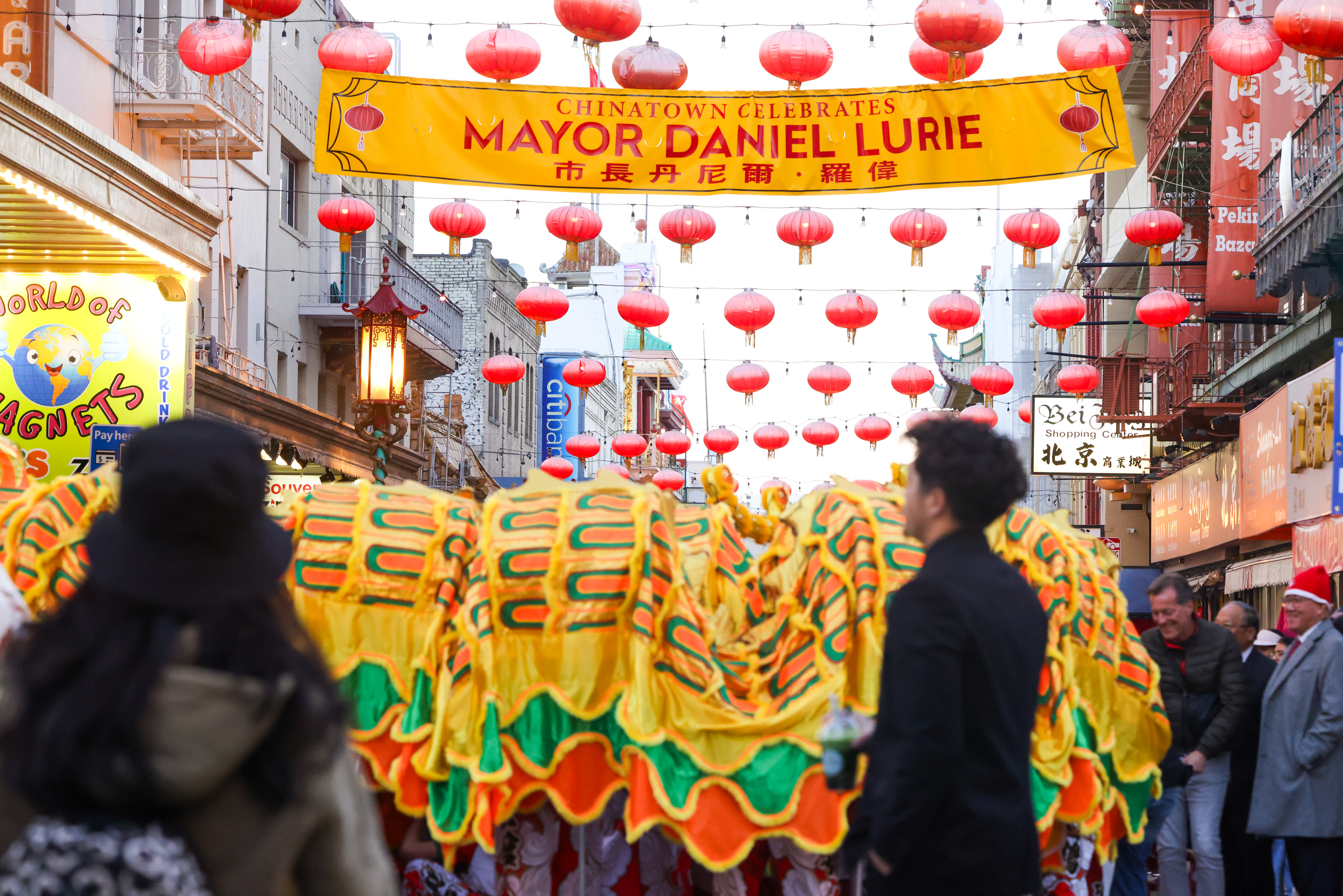 A vibrant street scene features a colorful dragon dance under red lanterns and a yellow banner celebrating Mayor Daniel Lurie in Chinatown.