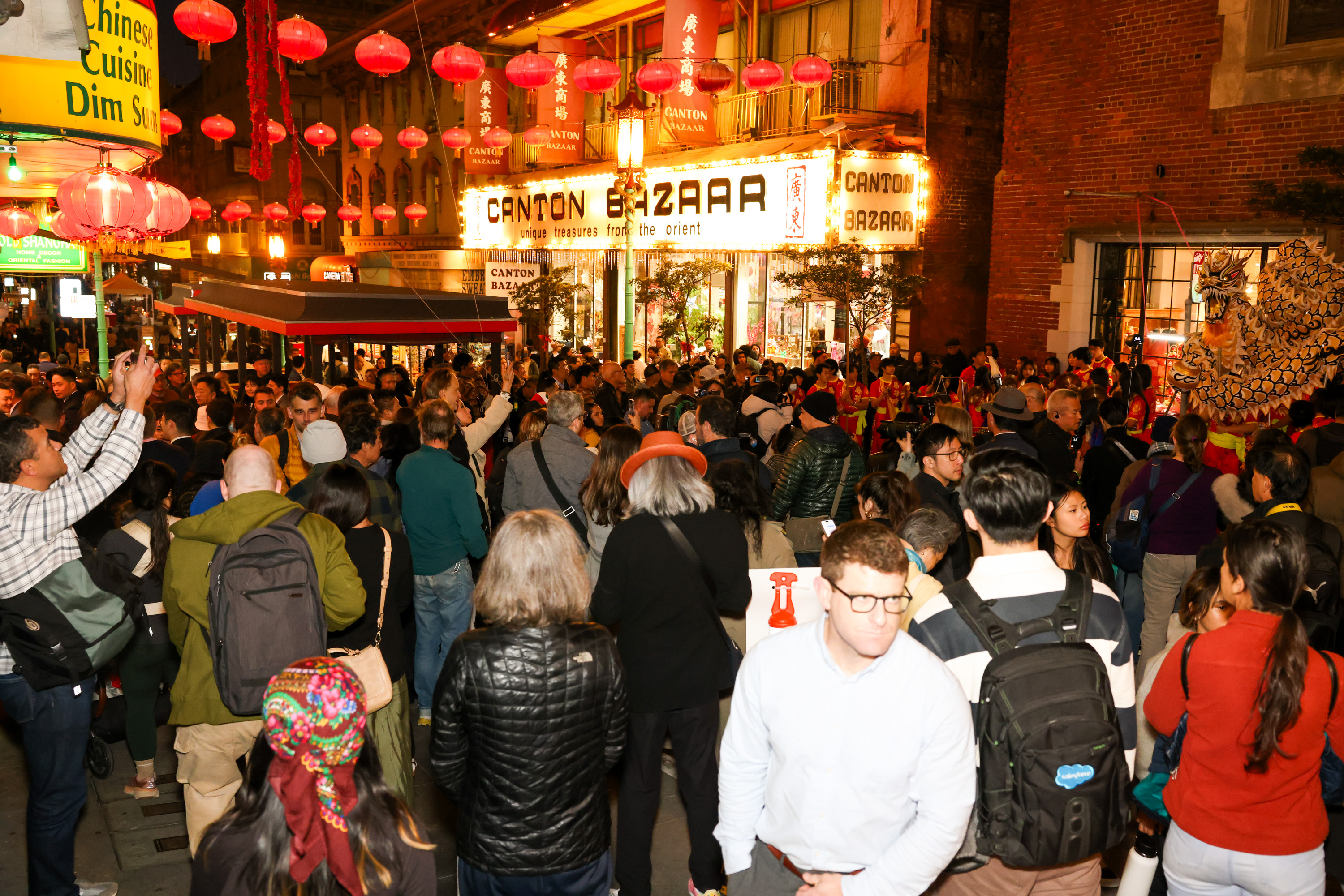 A crowded street scene with people gathered under red lanterns, near a store named &quot;Canton Bazaar.&quot; A dragon dance performance is taking place.