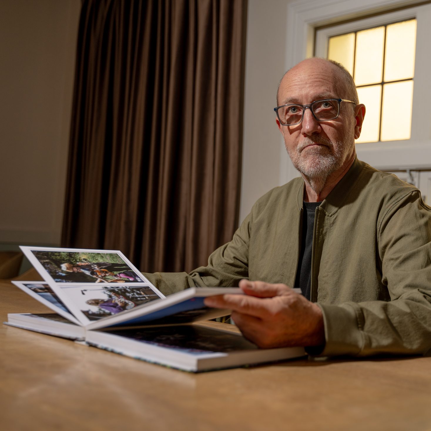 A man with glasses is sitting at a table, looking through a photo album. He wears a green jacket, and a window with patterned glass is behind him.