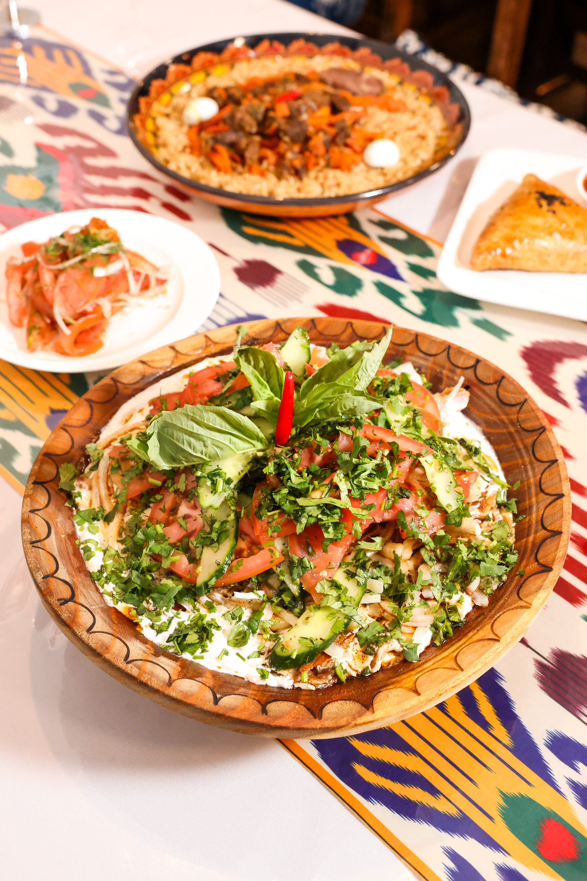 A vibrant meal with a central dish of fresh herbs, tomatoes, and cucumbers, accompanied by a plate of rice with meat and eggs, and a side of sliced vegetables.