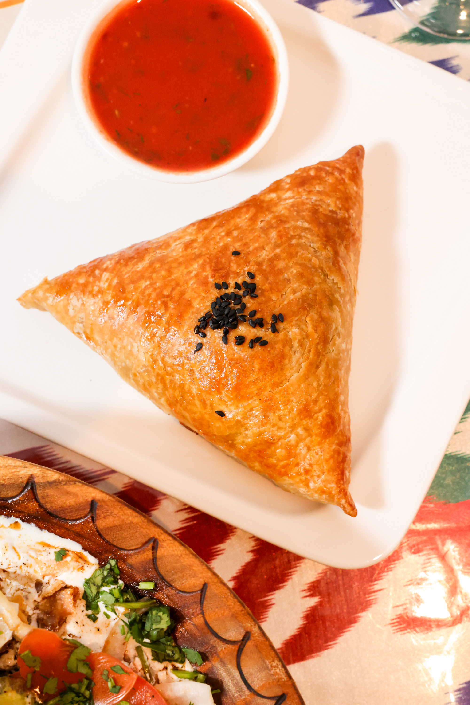 A golden triangular pastry with black seeds on top sits on a white plate beside a small bowl of red dipping sauce. Part of a salad is visible below.