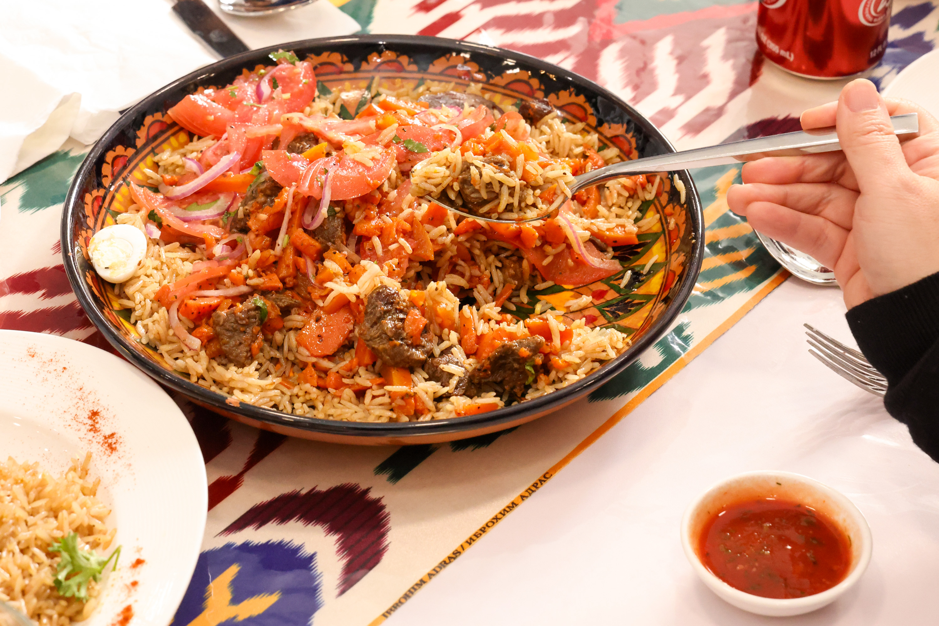 A colorful plate of rice with vegetables and meat, garnished with tomato slices and onions. A hand holds a spoon, and a small bowl of sauce is nearby.