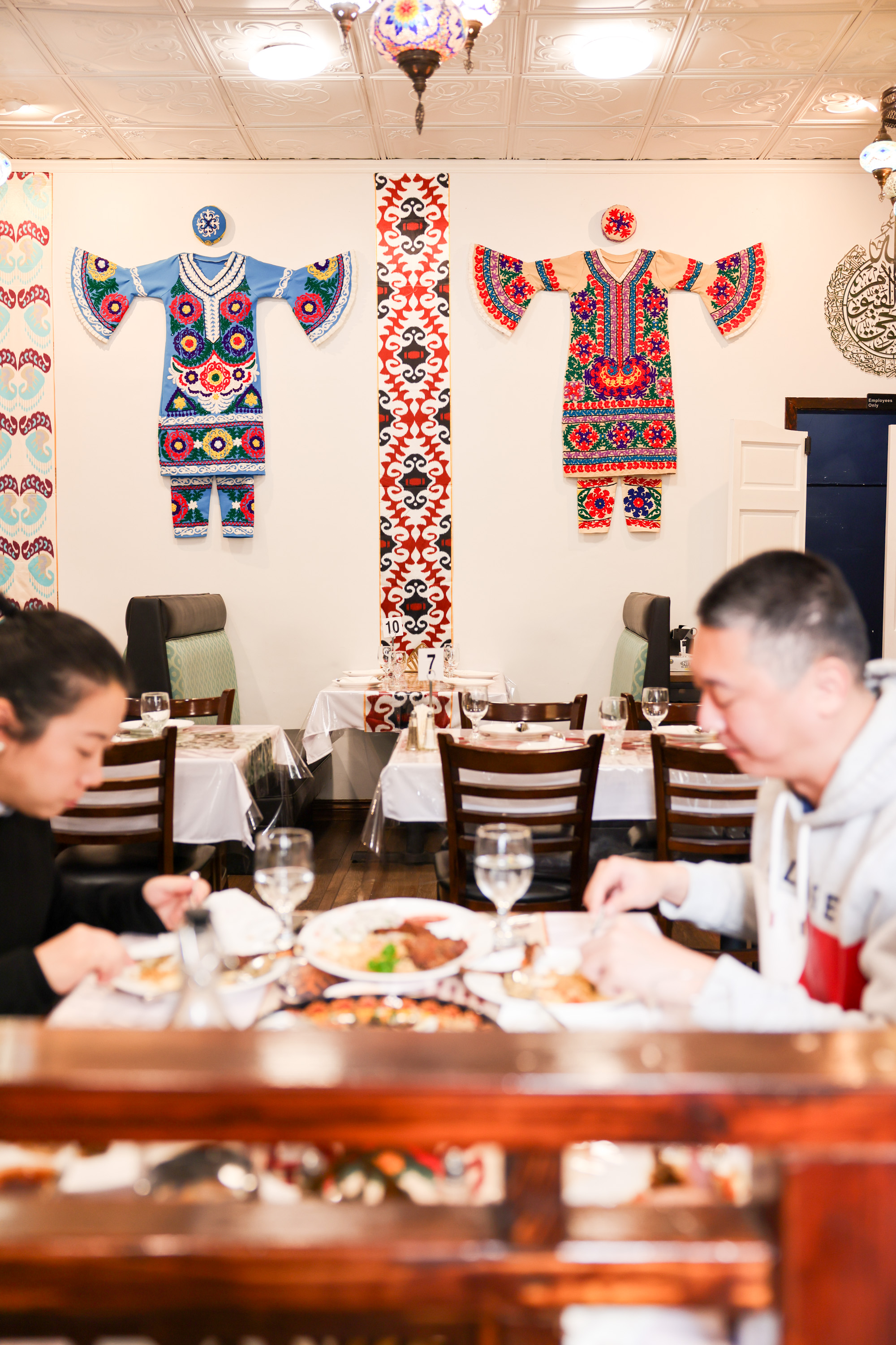 Two people dine in a restaurant with colorful, traditional patterned outfits mounted on the wall and numbered tables set with utensils and glasses.