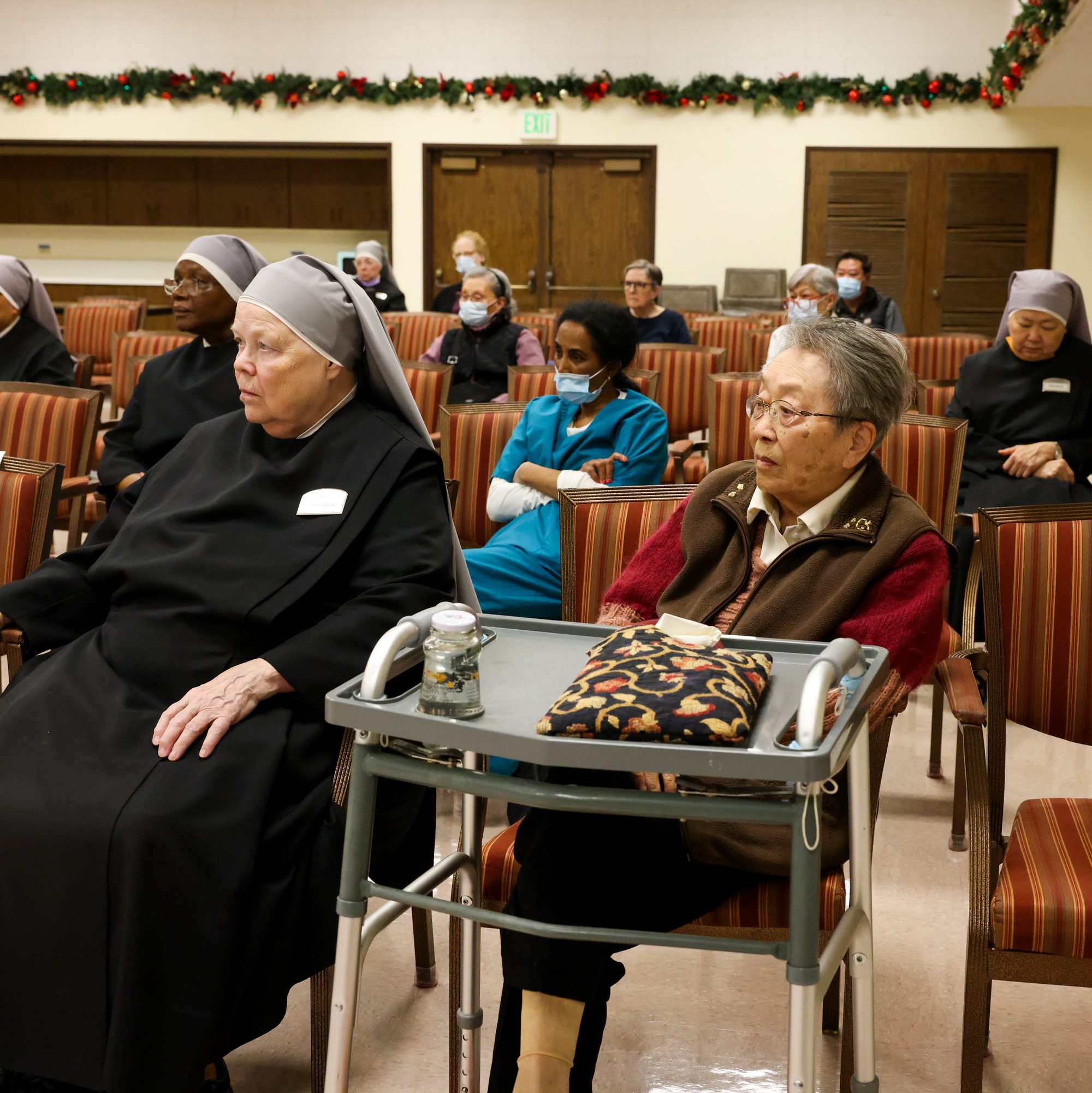 Elderly individuals, some wearing religious attire, sit on striped chairs in a room decorated with garlands. A sign reads &quot;Reserved,&quot; with a walker in front.