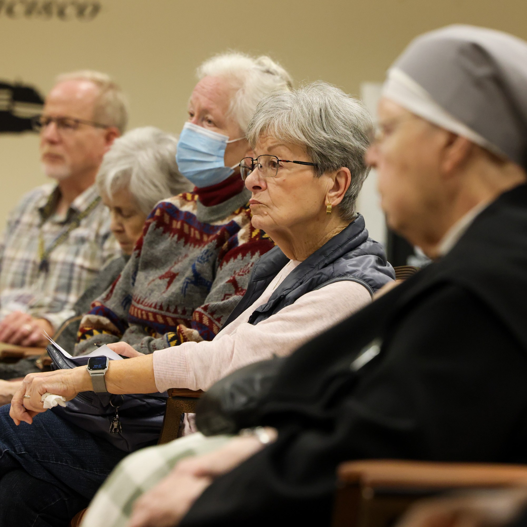 A group of elderly people, some wearing masks, sit attentively in a room. One person in a nun's habit is seen on the right. They appear to be listening intently.