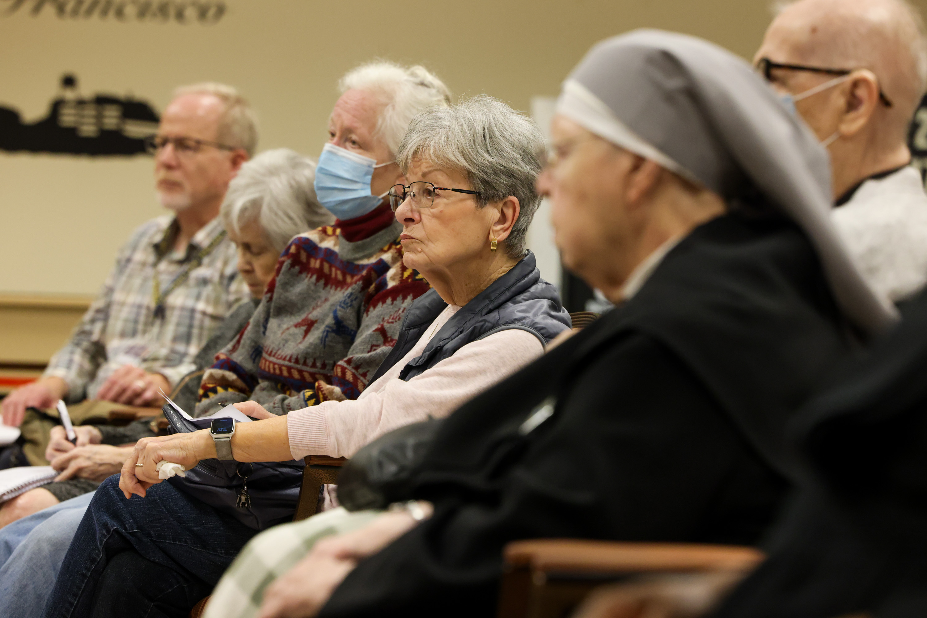 A group of elderly people, some wearing masks, sit attentively in a room. One person in a nun's habit is seen on the right. They appear to be listening intently.