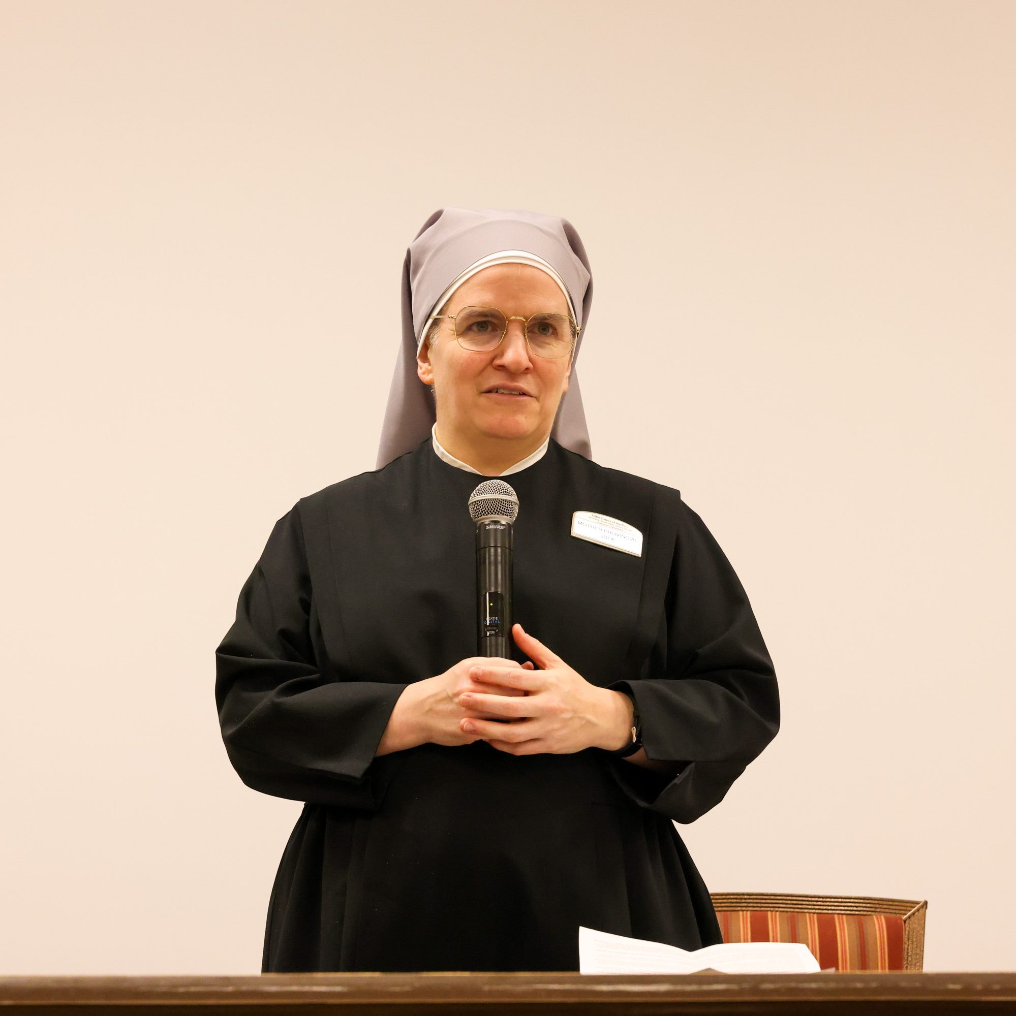 A woman in a black habit and gray veil speaks into a microphone. She stands behind a desk with papers, with two additional microphones on either side.