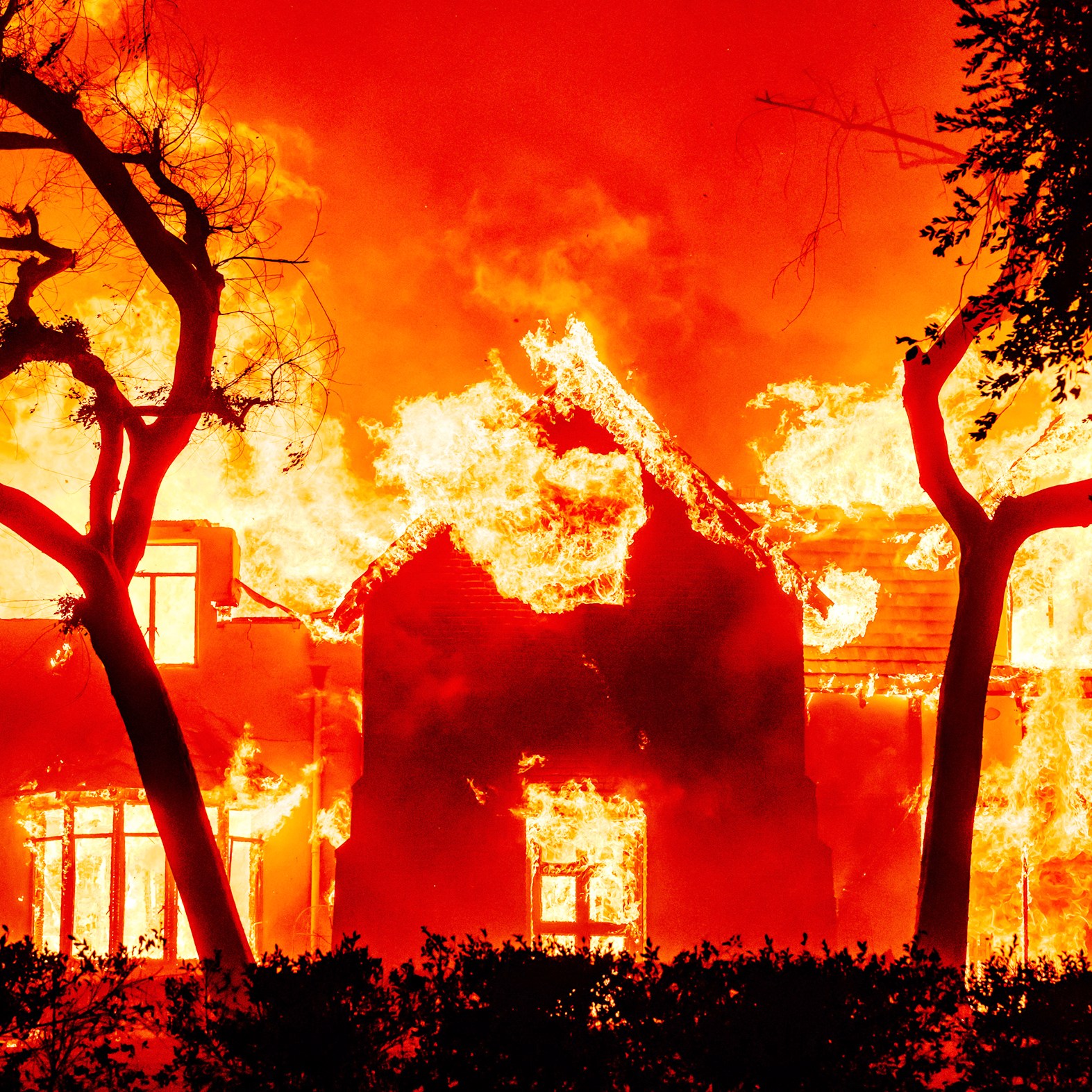 A house is engulfed in bright, intense flames amidst a fiery orange-red sky, with silhouetted trees in the foreground.