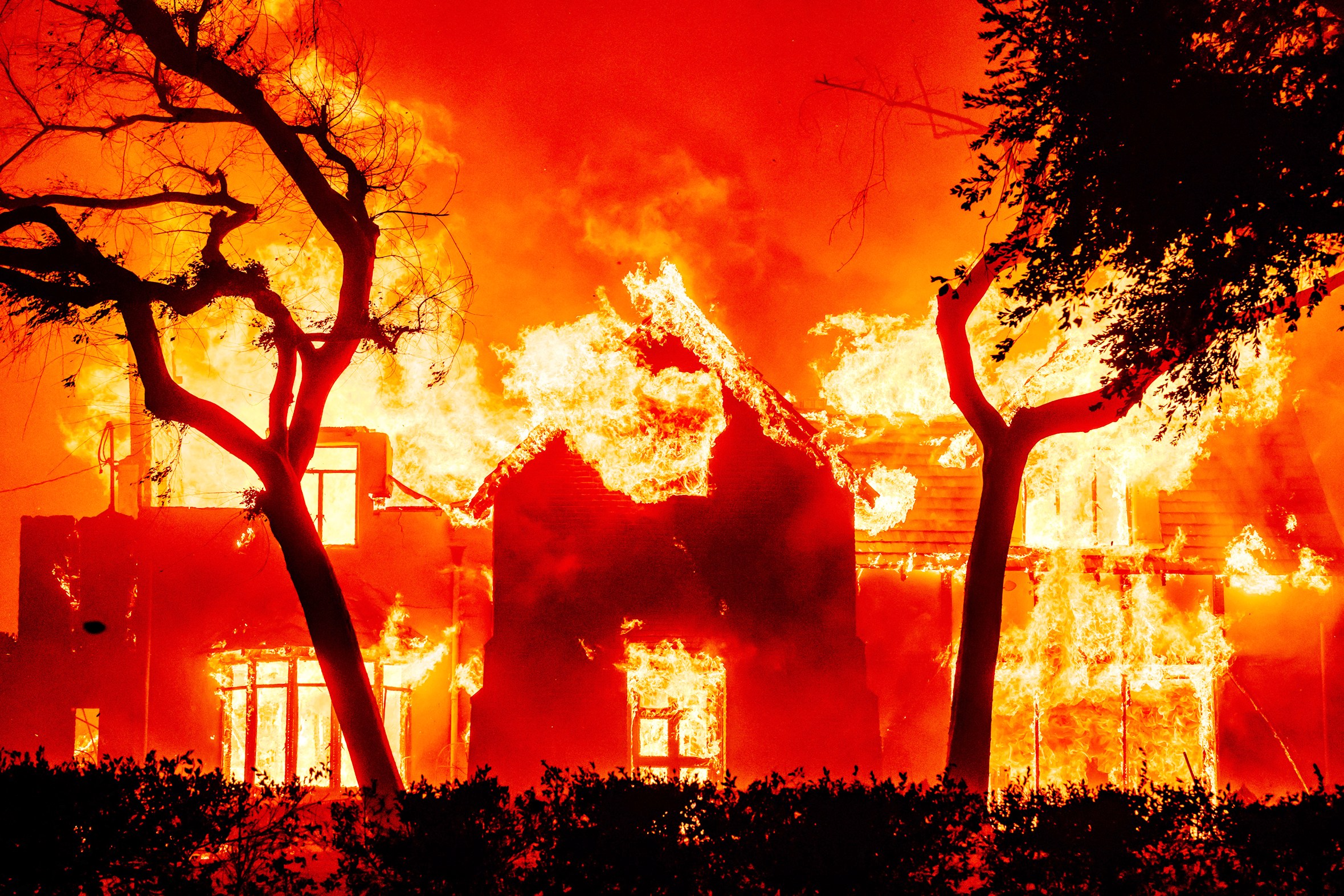 A house is engulfed in bright, intense flames amidst a fiery orange-red sky, with silhouetted trees in the foreground.