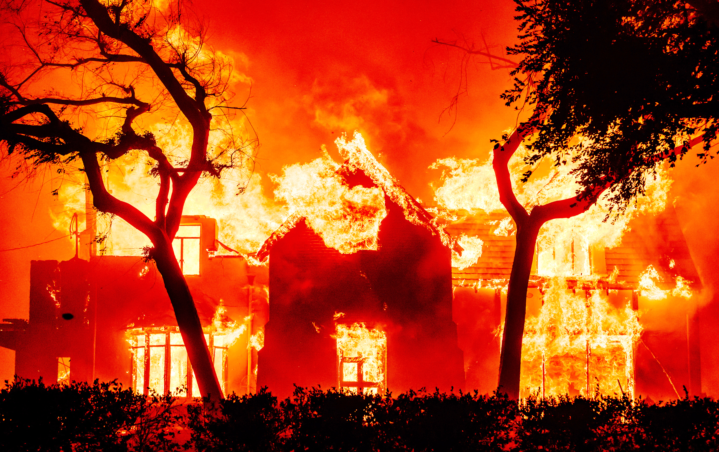 A house is engulfed in bright, intense flames amidst a fiery orange-red sky, with silhouetted trees in the foreground.