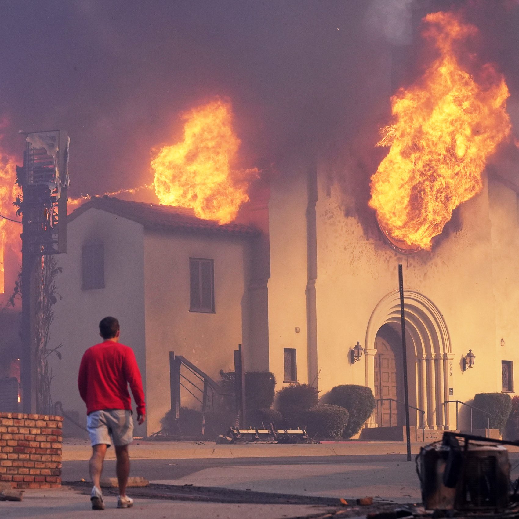 A building engulfed in large flames with smoke darkening the sky. A person in a red shirt and shorts walks toward the blaze, surrounded by debris.