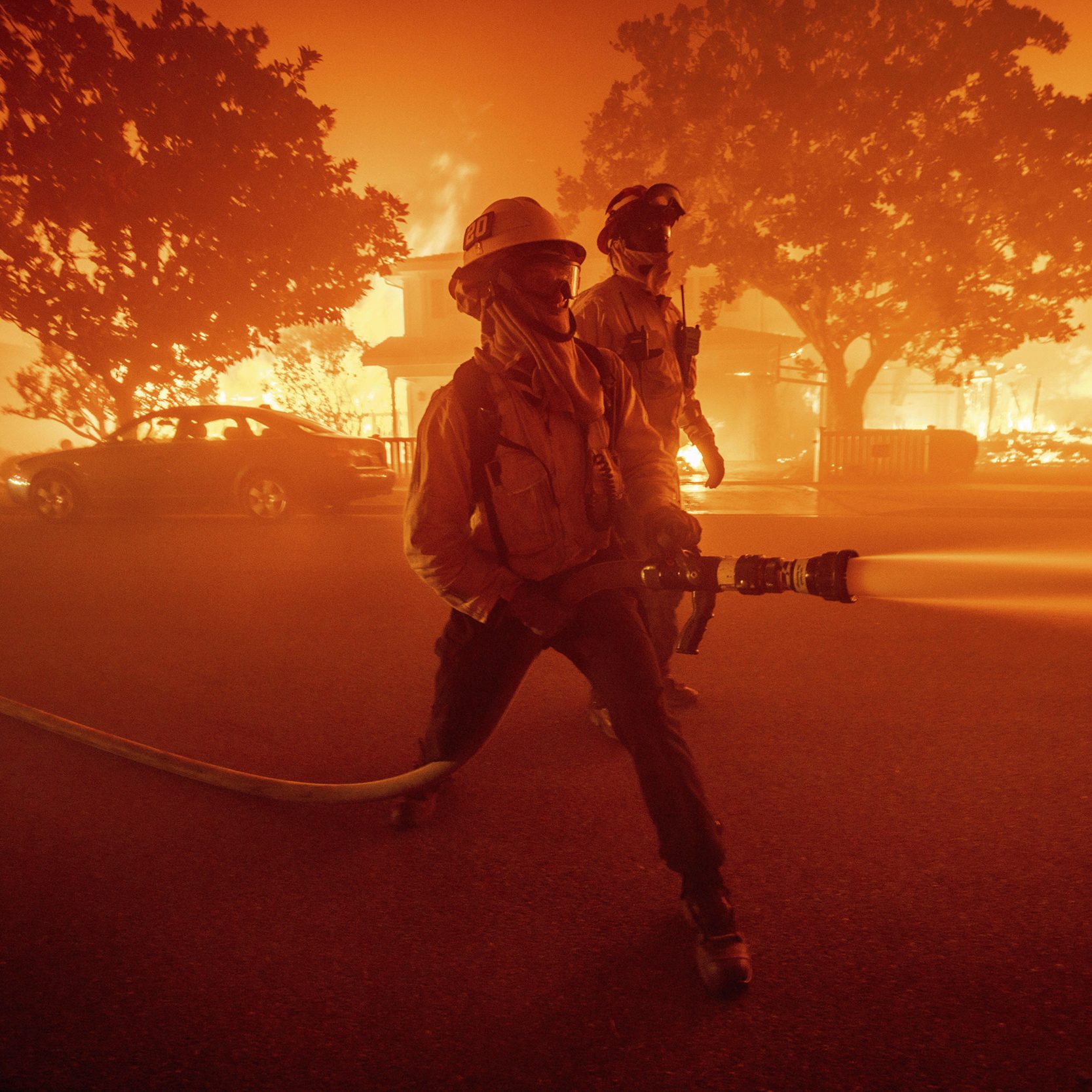 Two firefighters are battling a blaze in a street engulfed in fire, with burning buildings and trees, under an intense orange sky.