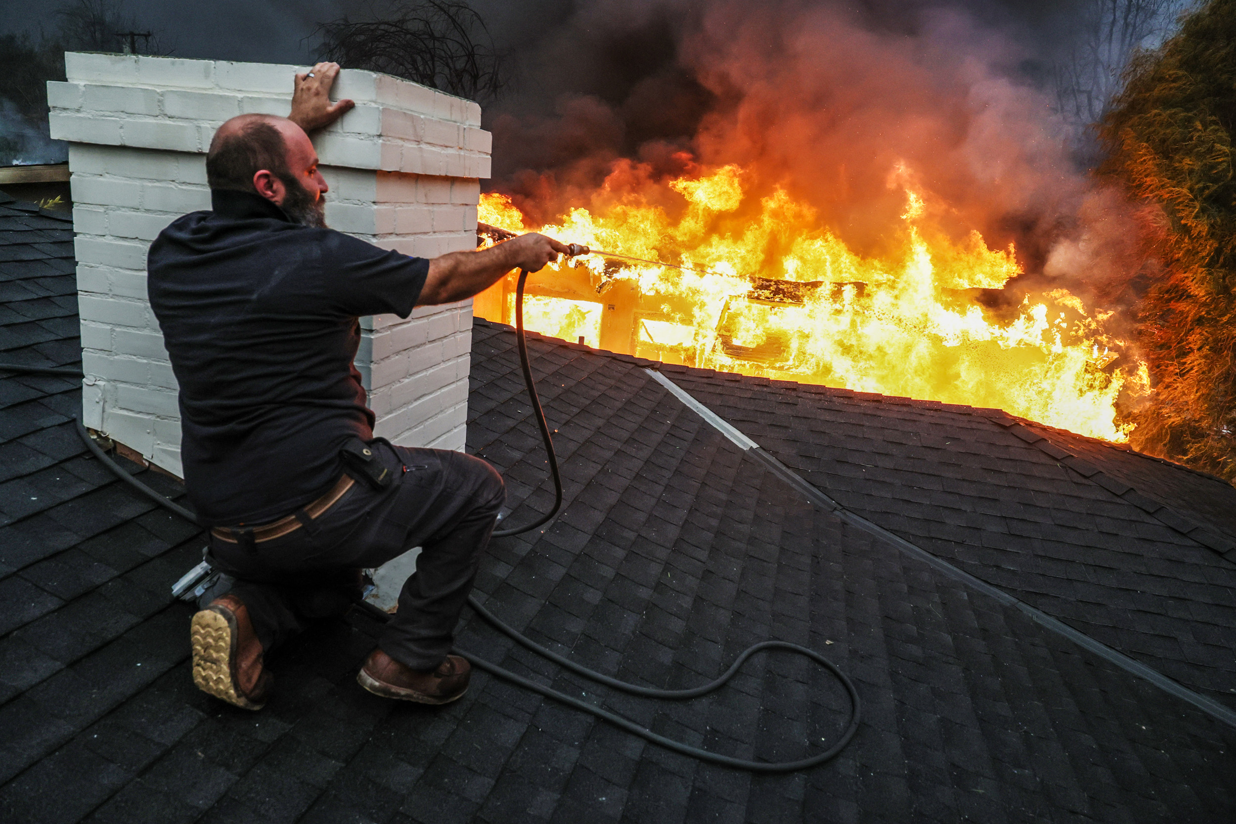 A person kneels on a rooftop with smoke and intense flames in the background, aiming a hose at the fire. The setting is urgent and dangerous.